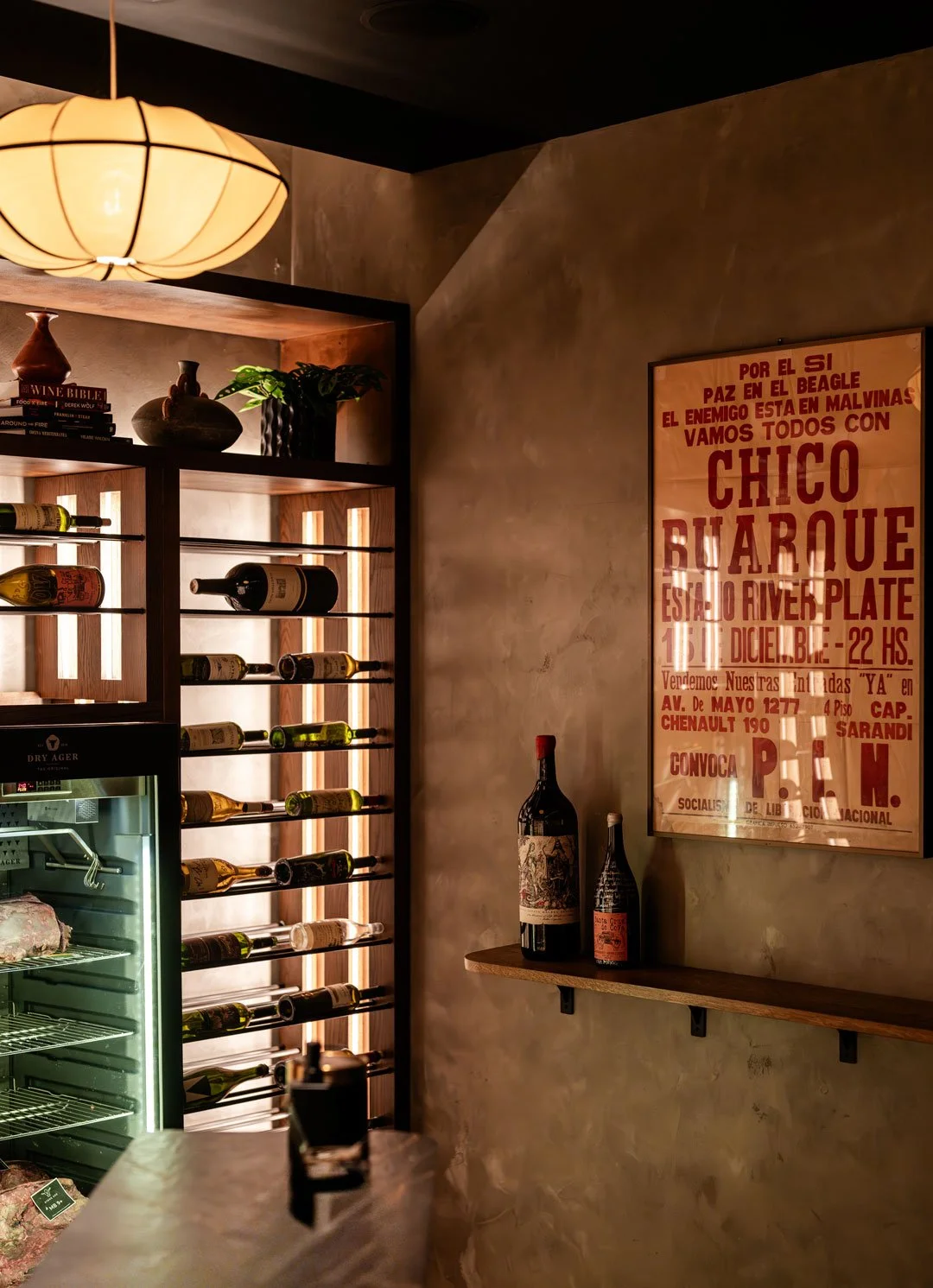 El Che wine display by Siren Betty Design with backlit wood shelving, a dry-aging case, warm plaster walls, and vintage Argentine artwork.