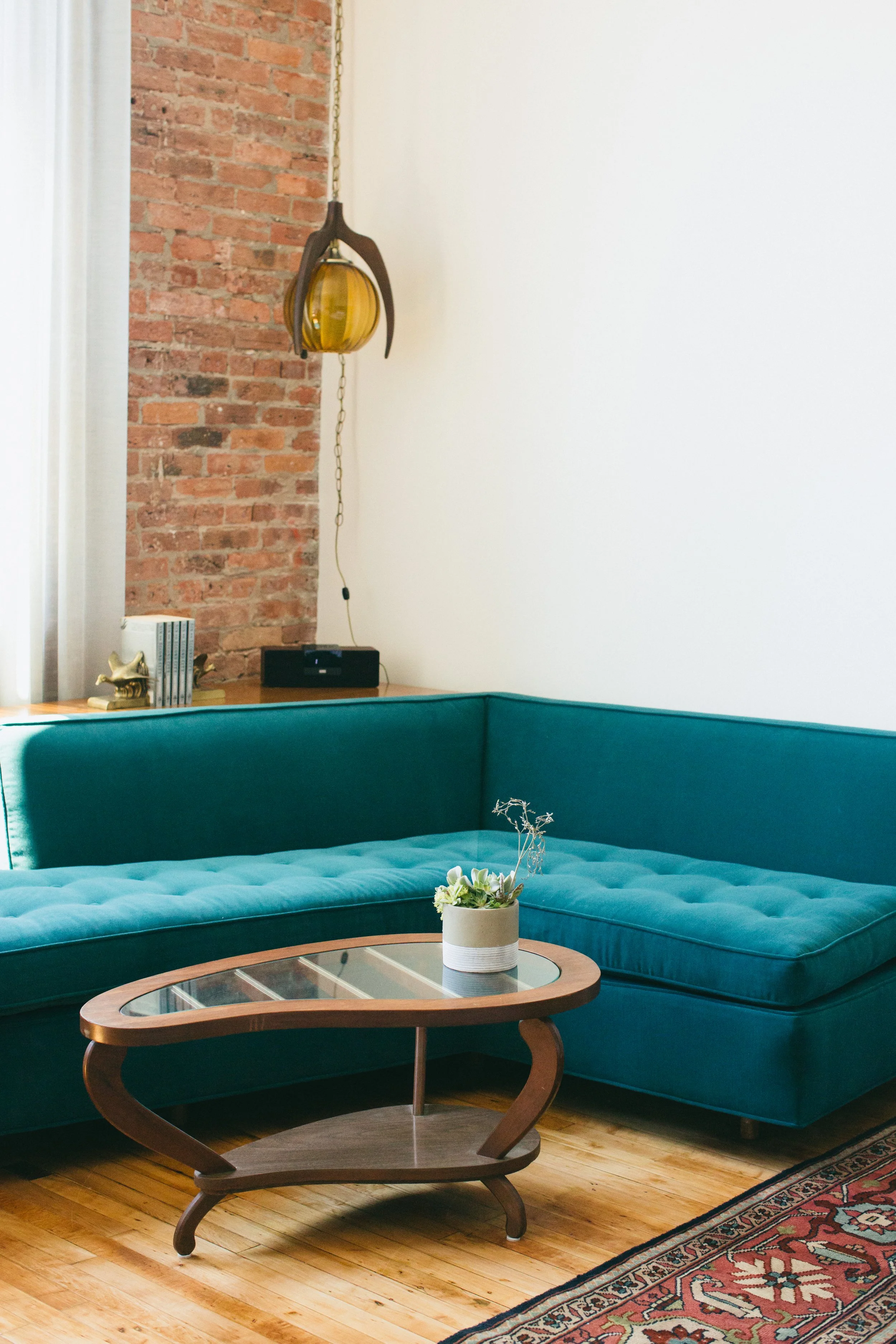 The Publishing House by Siren Betty Design, teal mid-century sectional with amber pendant light and exposed brick wall.