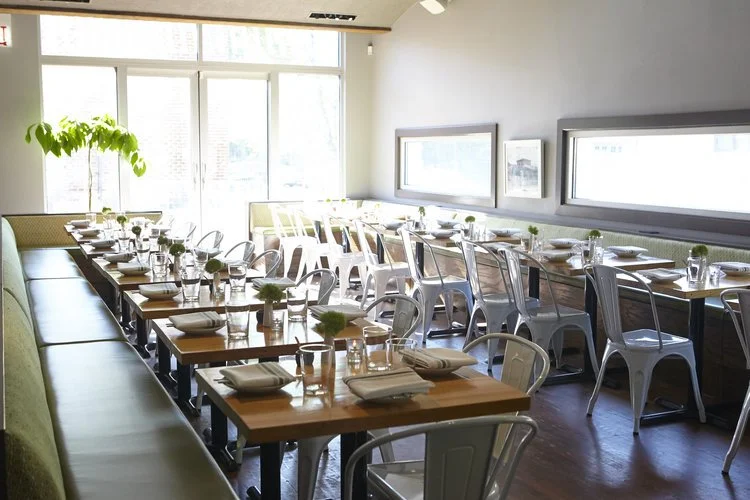 Quiote by Siren Betty Design. Sunlit dining with light wood tables, white metal chairs, and a long green banquette.