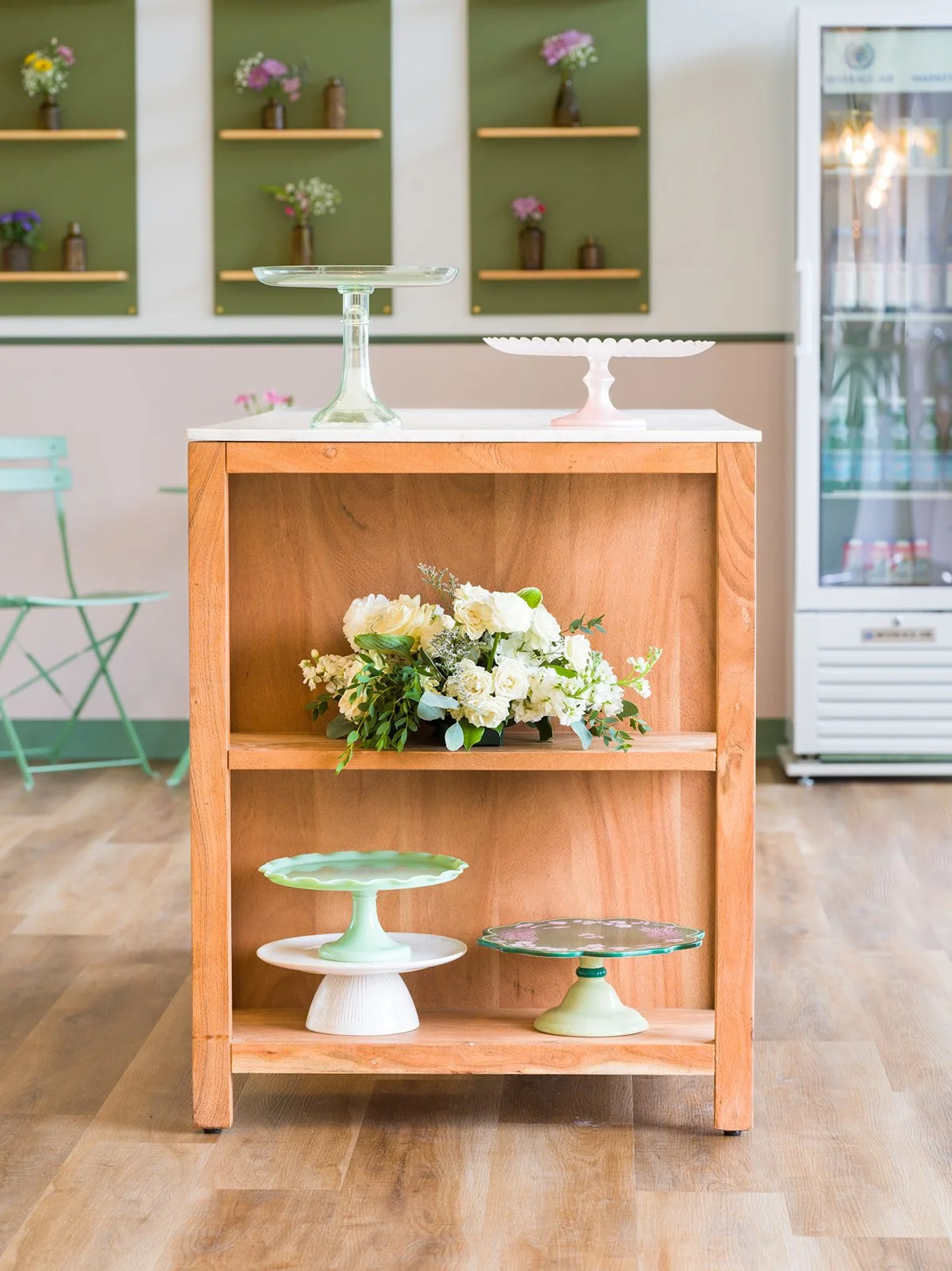 Wood display cart with pastel cake stands and fresh florals at Après Pastry & Bakery by Siren Betty Design; arched shelves.