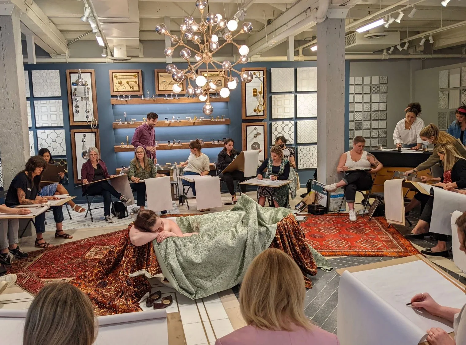 Life drawing session with artists sketching a reclining model in a studio filled with lighting displays and material samples.