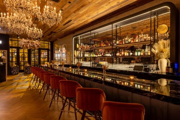 Hide & Seek bar by Siren Betty Design featuring black marble countertops, crystal chandeliers, and amber velvet bar stools in warm lighting.