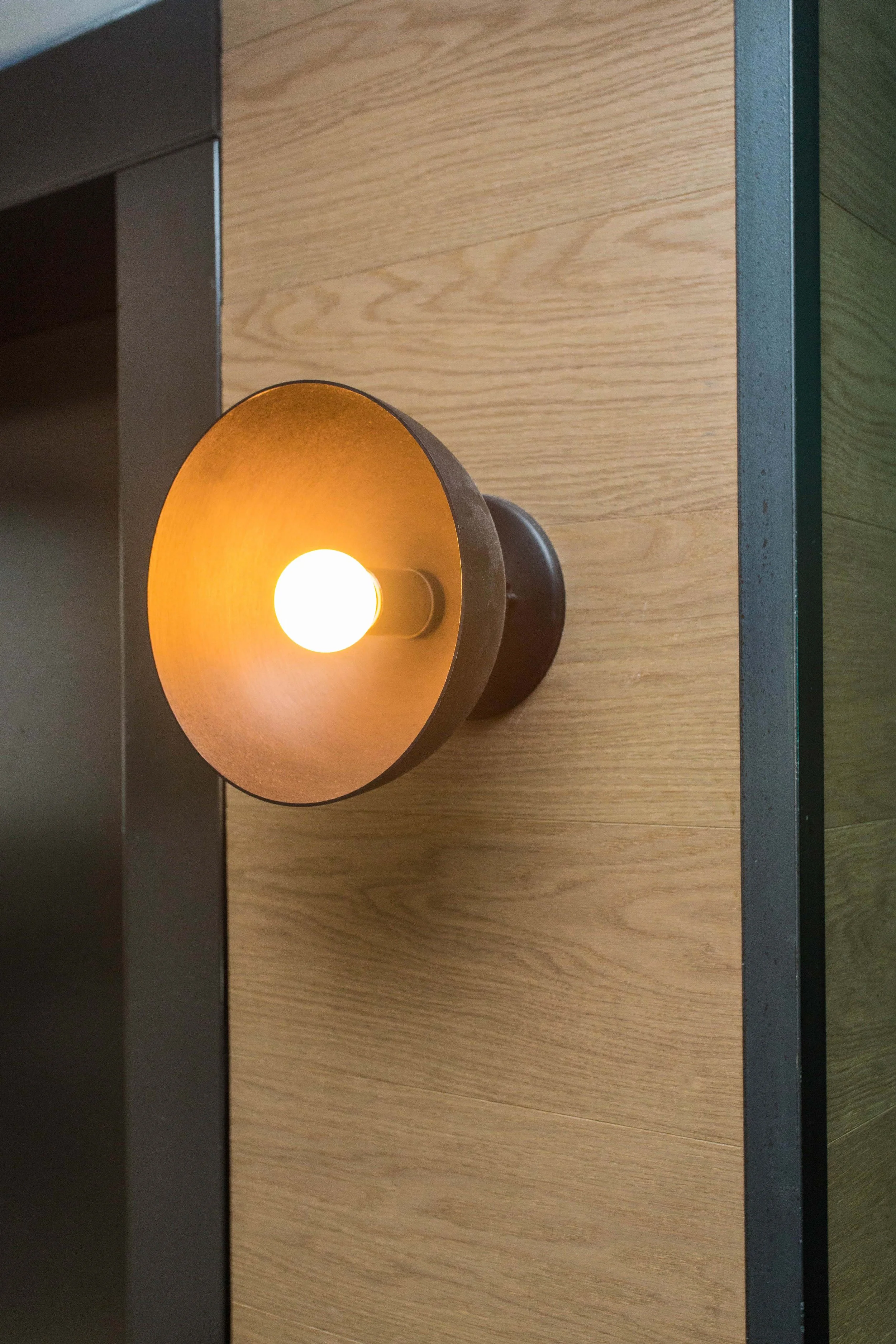 Benchmark by Siren Betty Design, warm wall sconce with brass finish mounted on light wood panel for ambient glow.