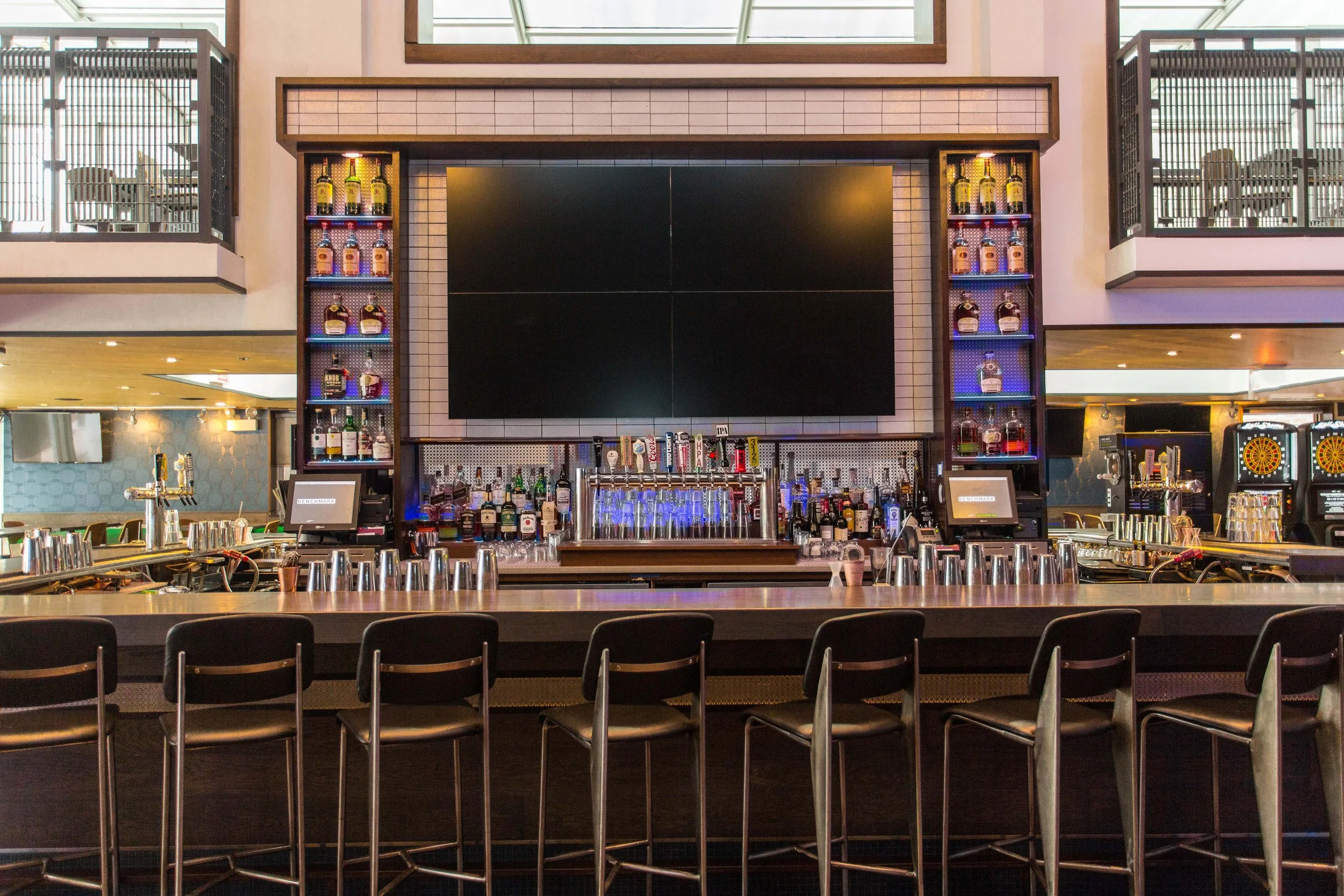 Benchmark by Siren Betty Design, modern hospitality bar with geometric tile backdrop, large screens, and backlit shelving.