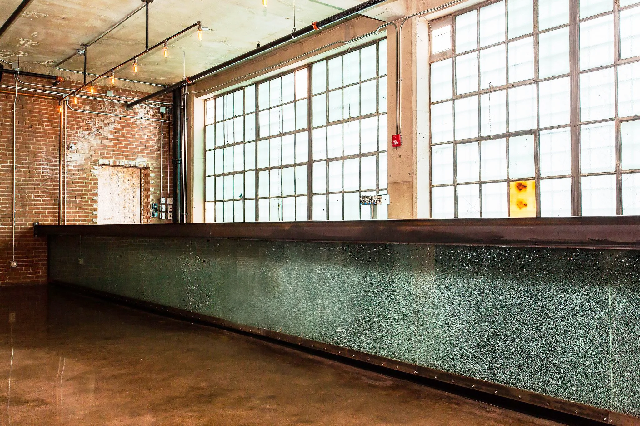Morgan MFG by Siren Betty Design. Long bar with patinated metal front, brick walls, glass block windows, and pendant bulbs.