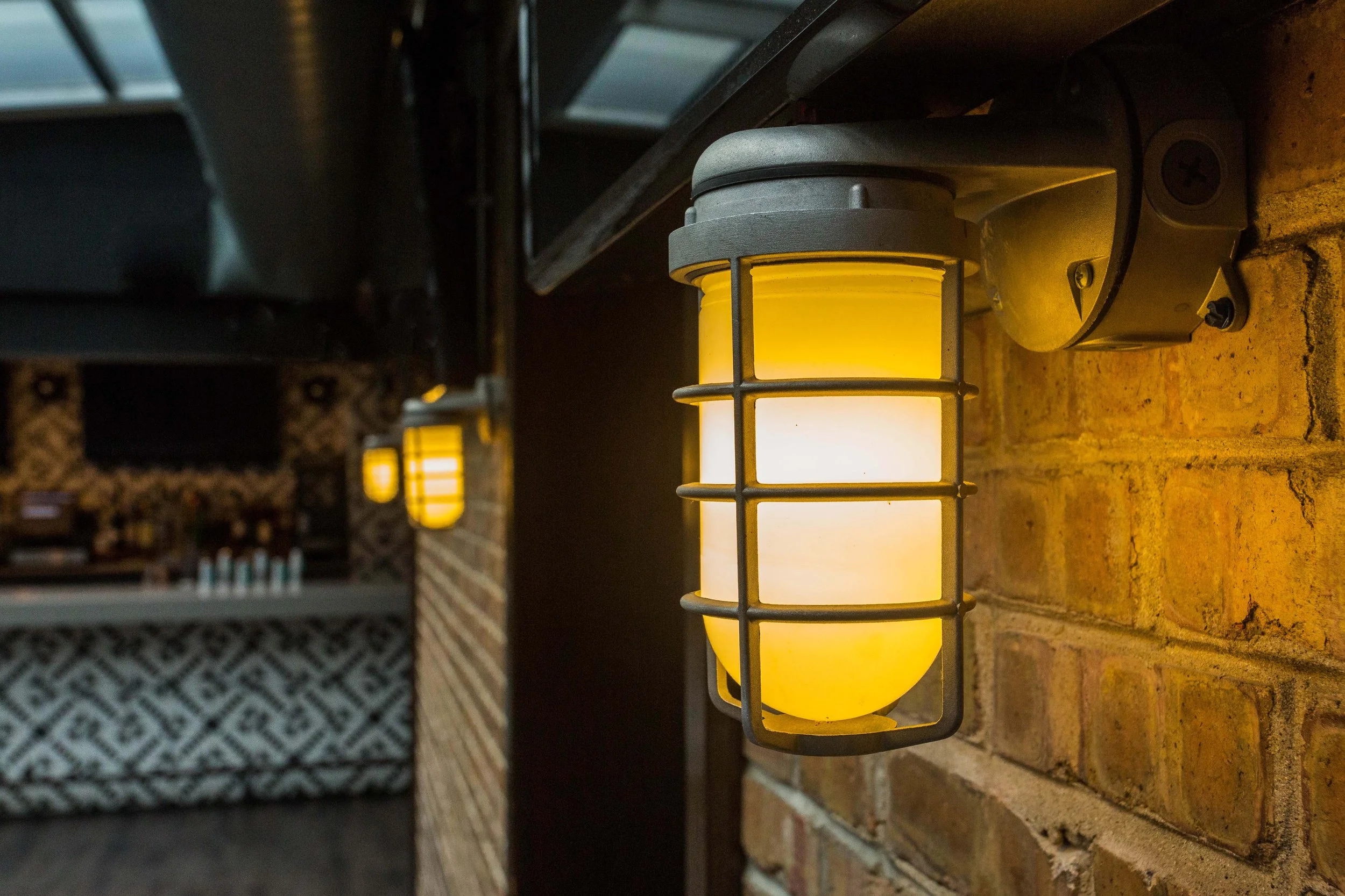 Benchmark by Siren Betty Design, close-up of warm industrial wall sconce illuminating brick walls near the bar area.