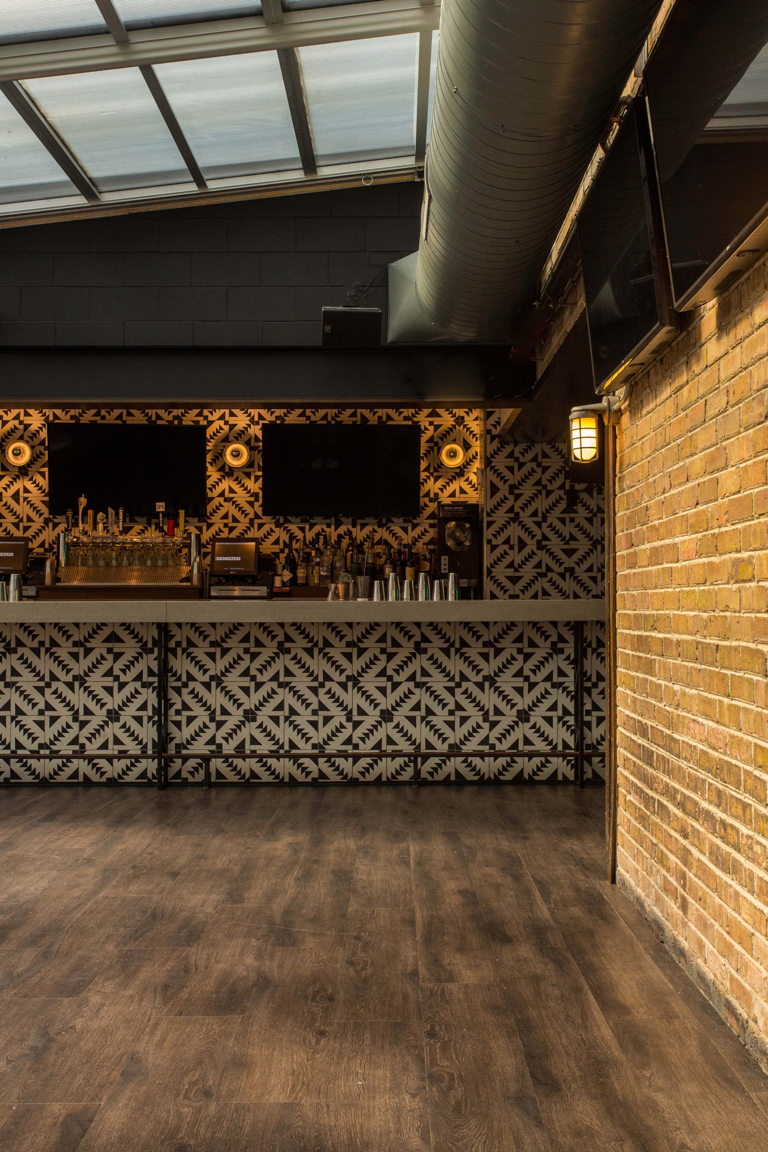 Benchmark by Siren Betty Design, patterned tile bar with warm sconces and exposed brick wall under skylit ceiling.
