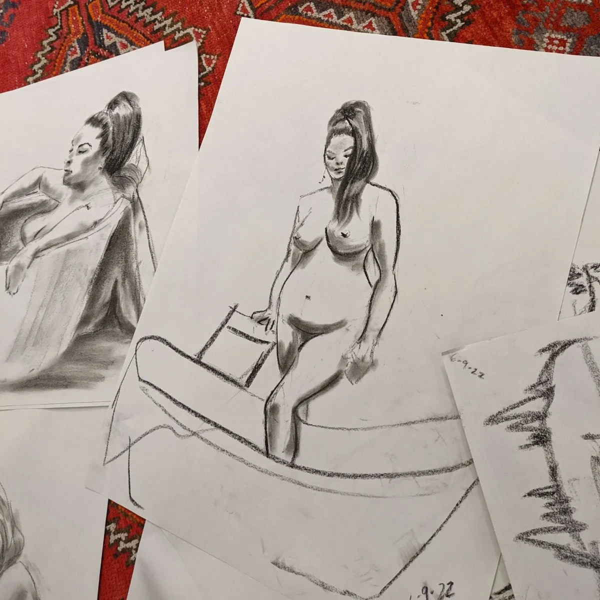 Charcoal life drawing sketches of a reclining and seated model displayed on paper over a patterned rug.