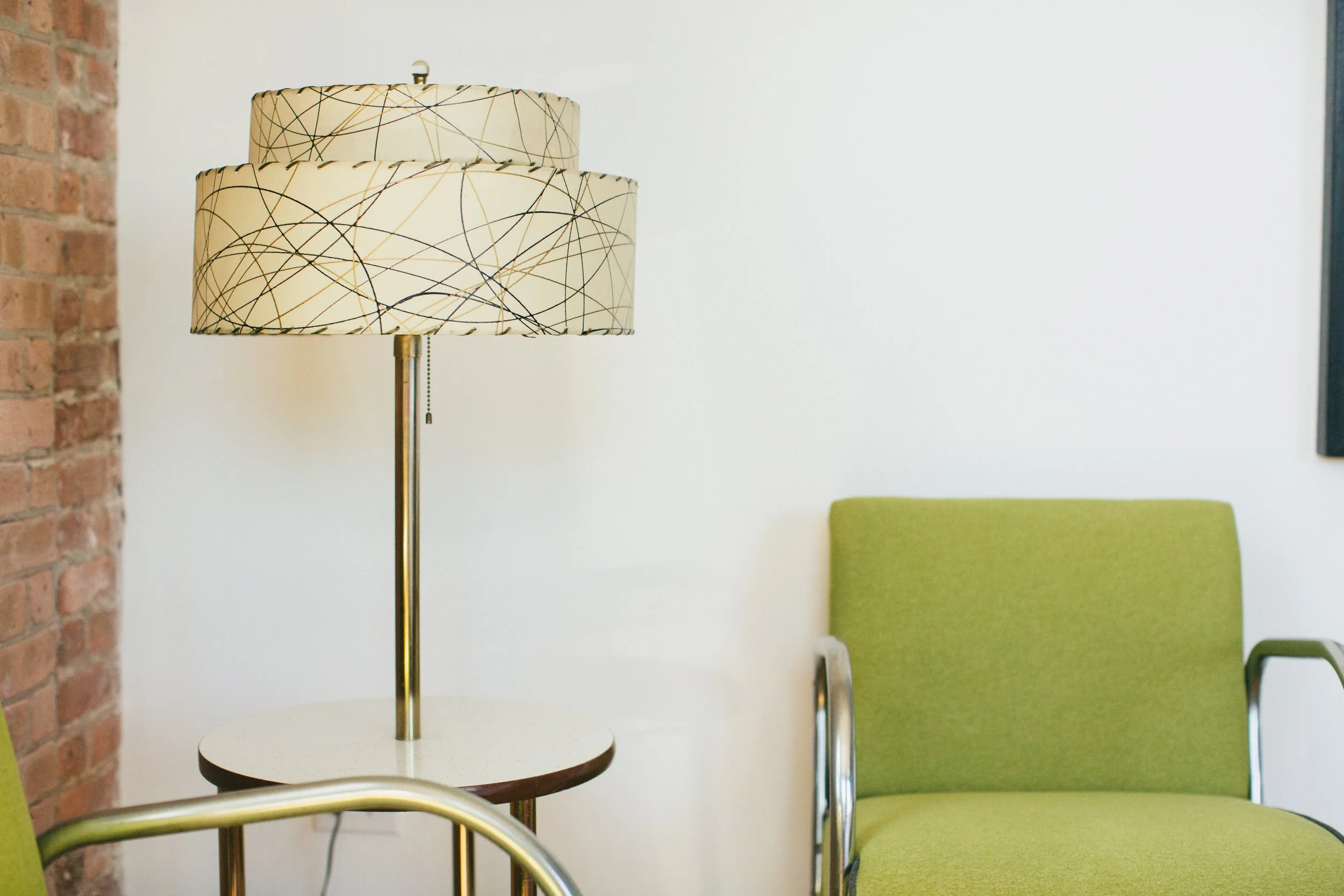 The Publishing House by Siren Betty Design, mid-century lamp with patterned shade beside green upholstered chair.