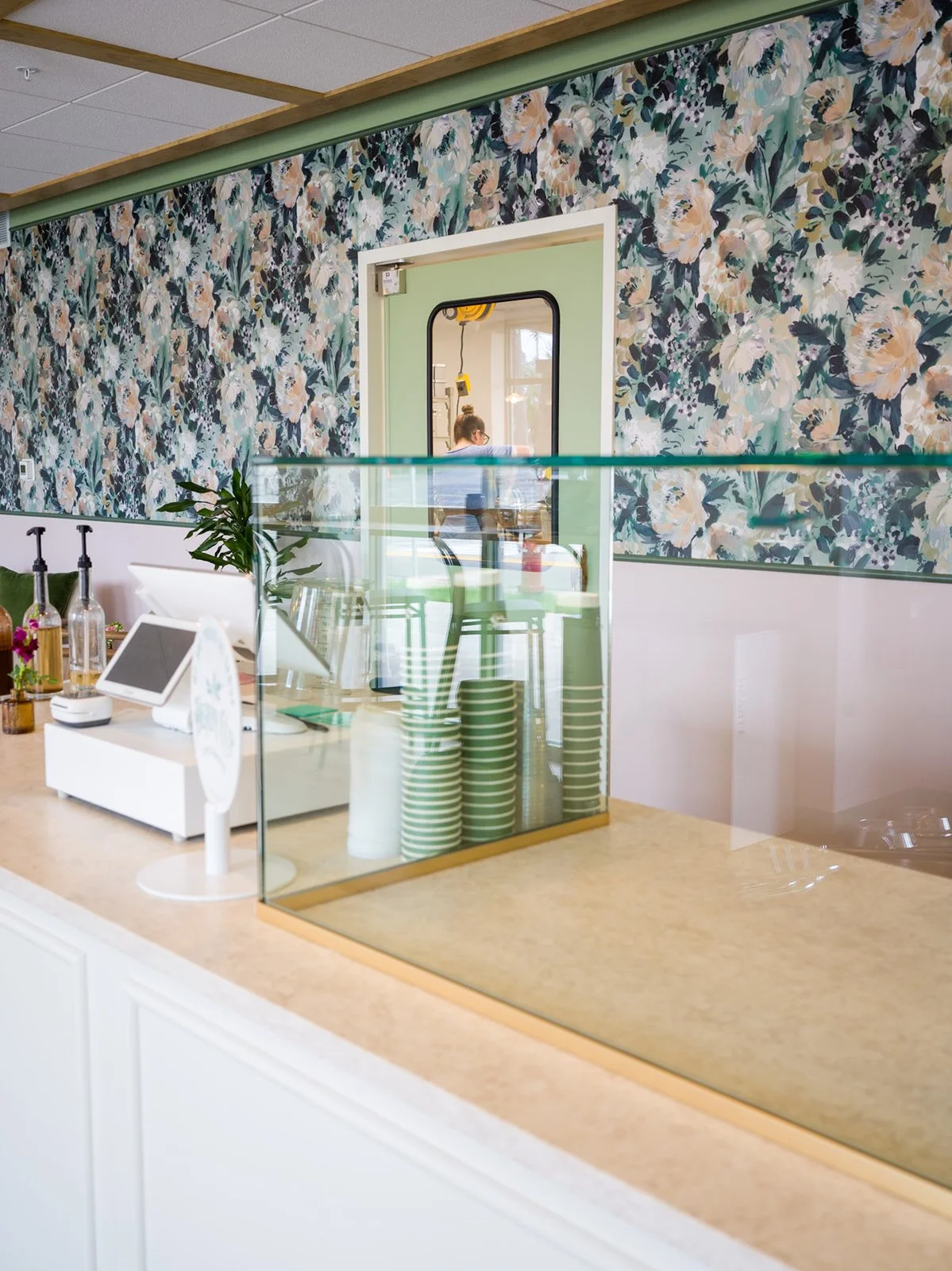 Service counter with glass guard, POS, and stacked striped cups before floral wall at Après Pastry & Bakery by Siren Betty Design.