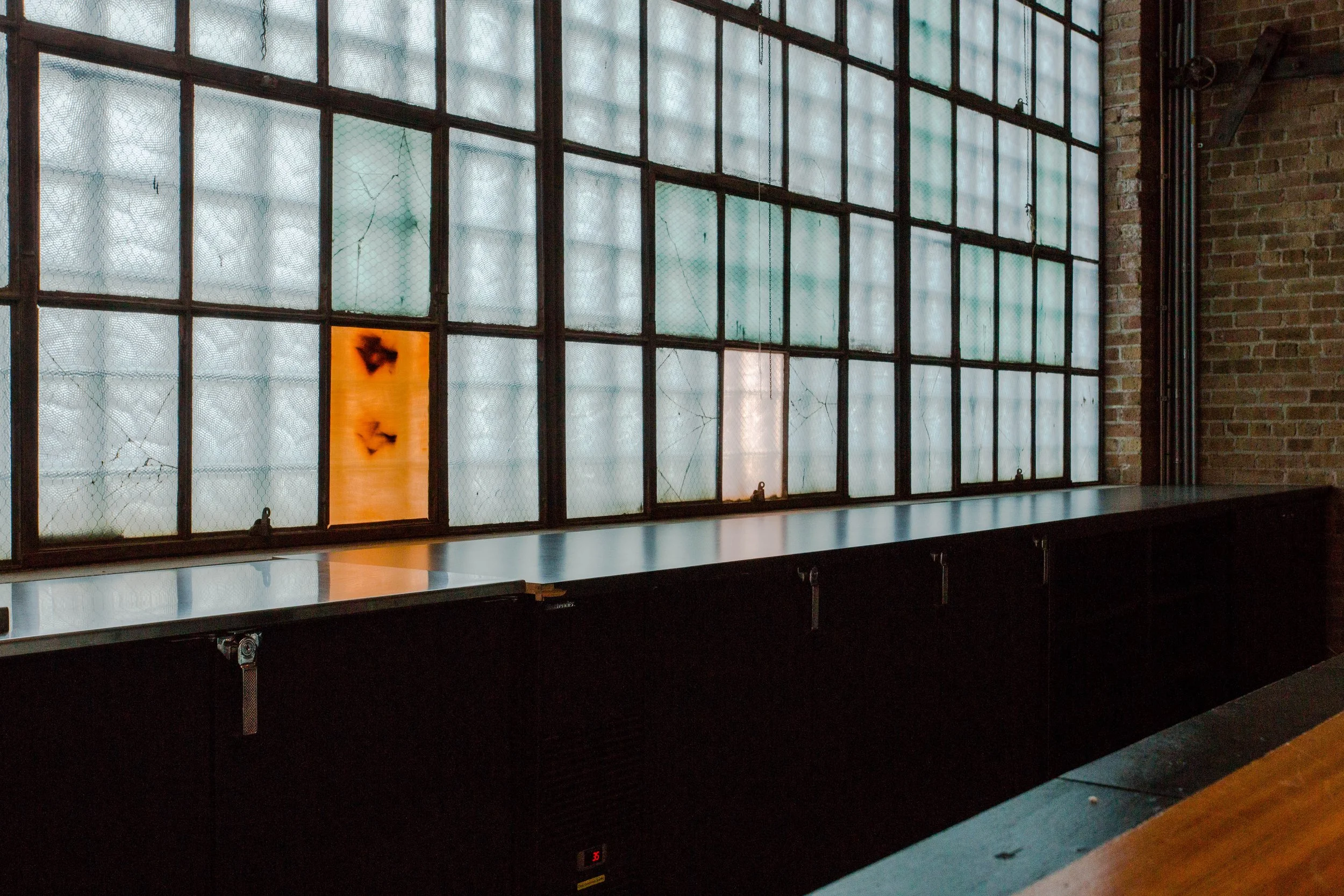 Morgan MFG by Siren Betty Design. Industrial bar back set against tall glass block windows with warm accent panels.