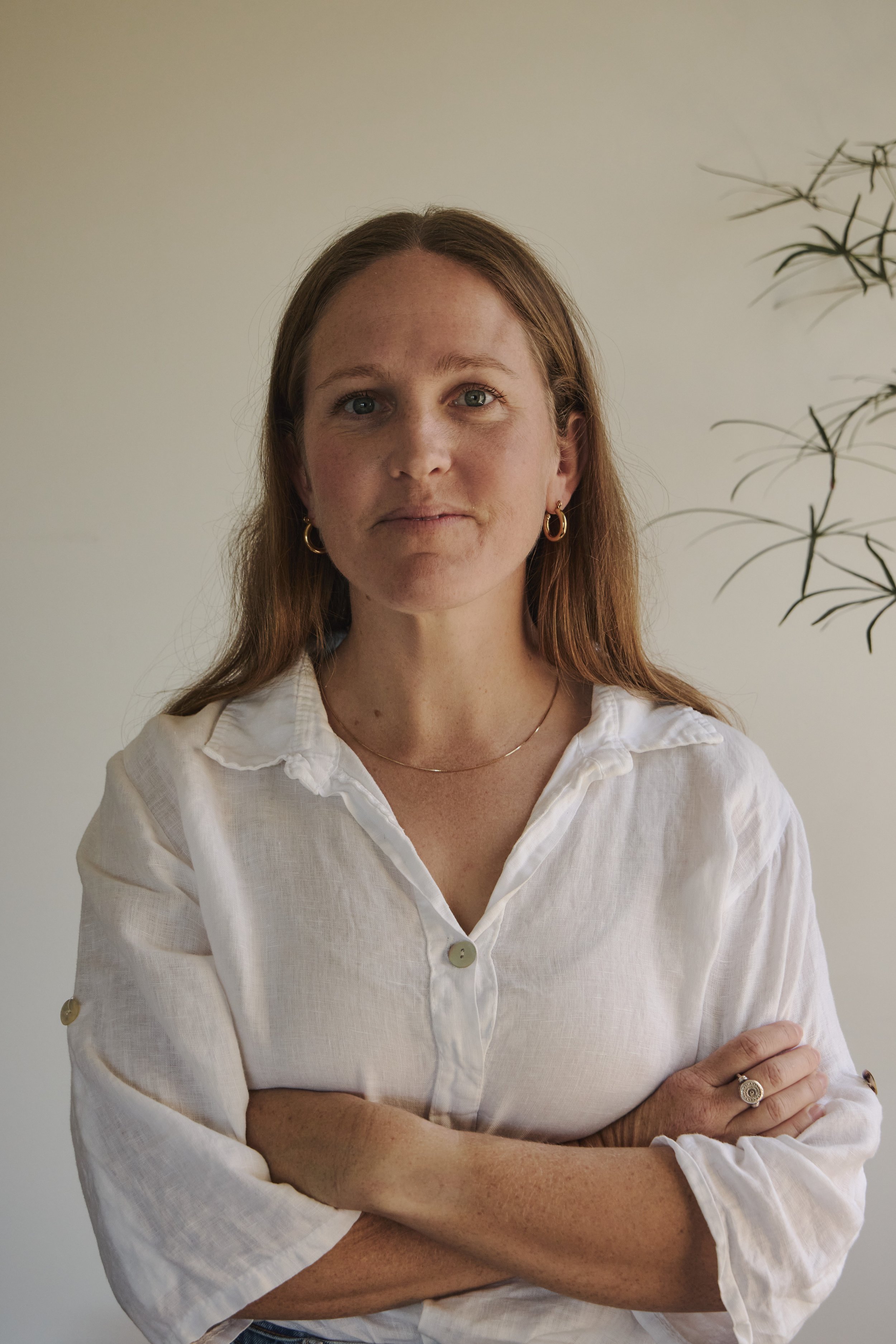 A woman with light brown hair, wearing a white button-up shirt, standing with arms crossed in front of a plain light-colored wall and a plant.