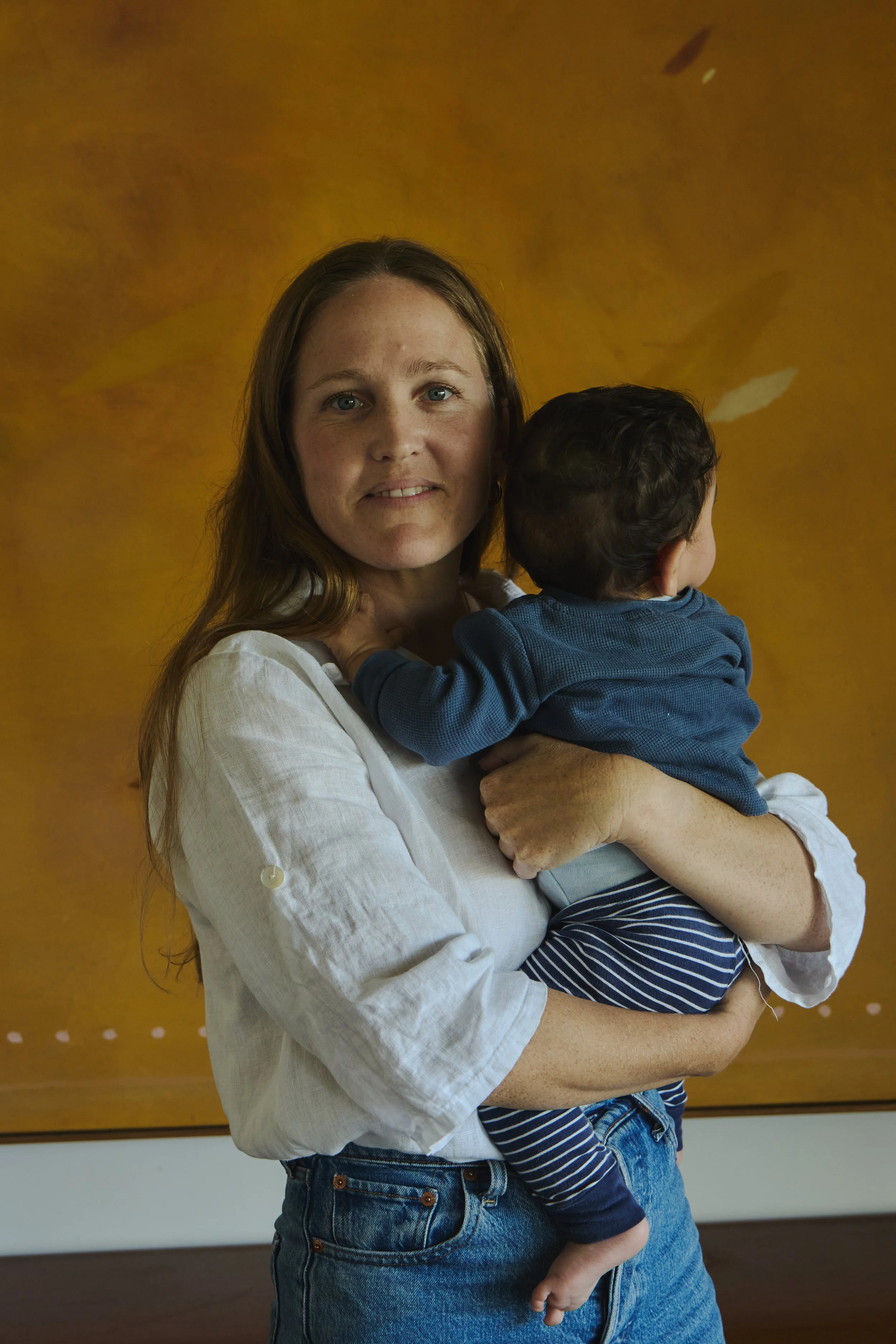 A woman with long hair holding a young child with dark curly hair against a yellow backdrop.