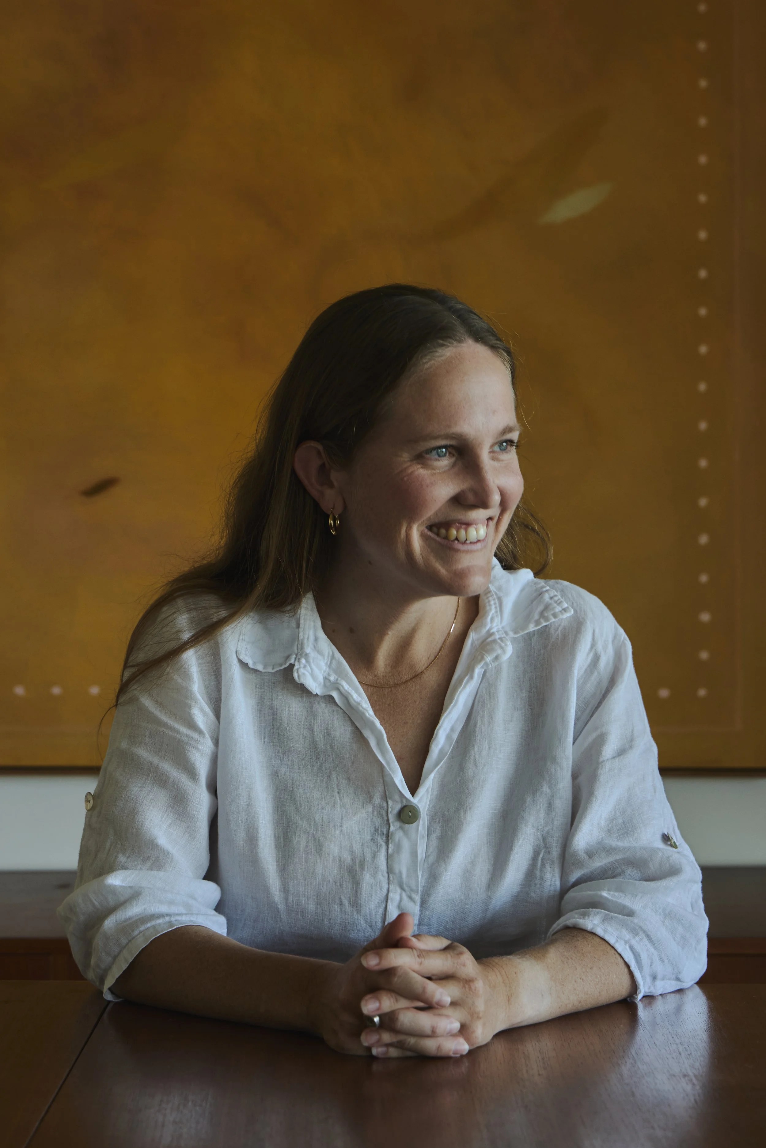 A woman with long brown hair, earrings, and a white shirt sitting at a wooden table, smiling while looking to the right. Behind her, there is a wooden wall with an abstract pattern.