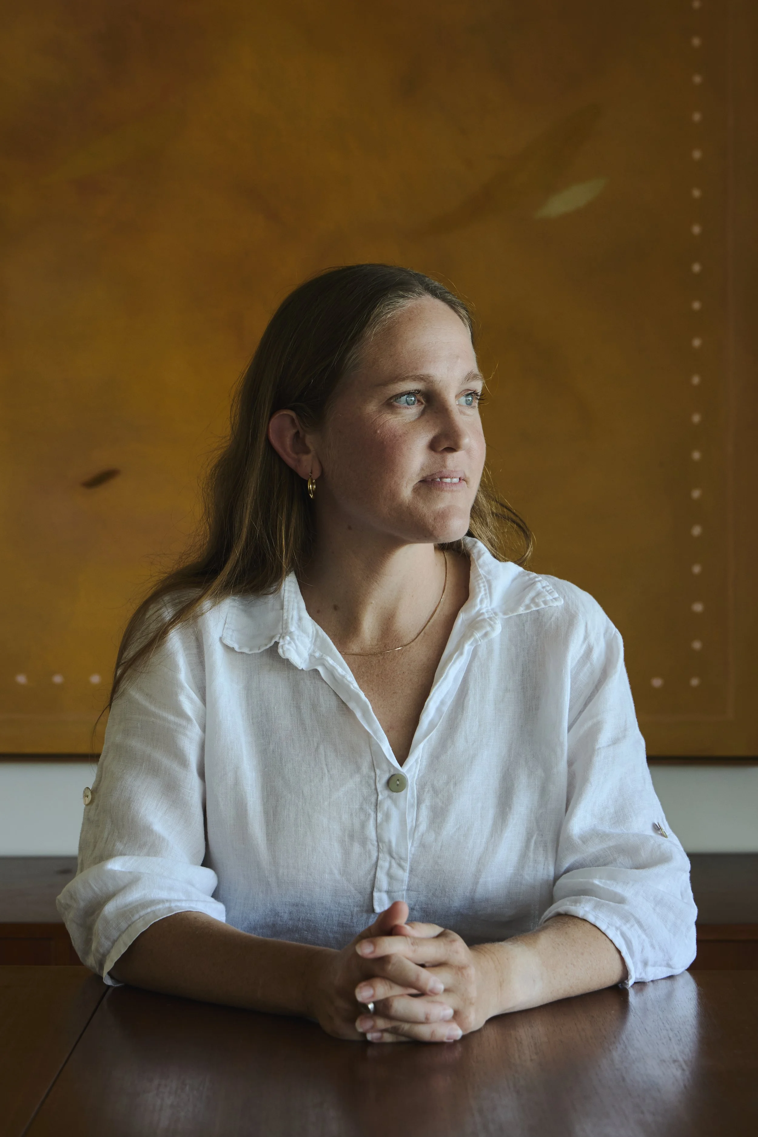 A woman with light brown hair wearing a white button-up shirt sits at a wooden table in front of a large abstract painting with warm colors. She has her hands clasped on the table and is looking slightly to her left.