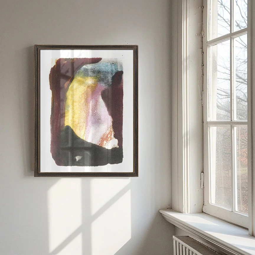 Refuge by Melissa McGill displayed in a dark wood frame beside a sunlit window, with burgundy and charcoal forms enclosing a luminous yellow and pale pink interior.