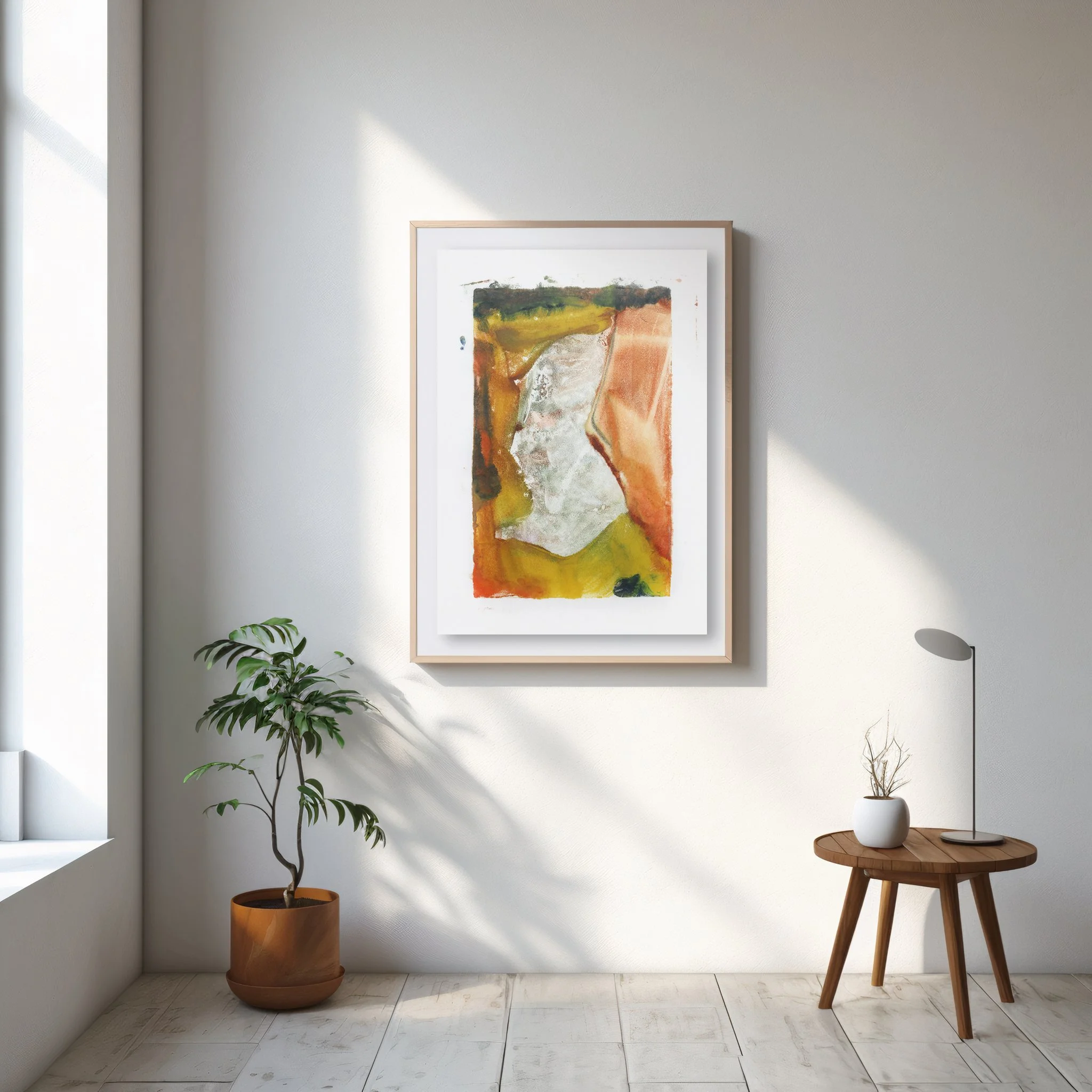 Floodplain by Melissa McGill displayed in a natural wood frame on a sunlit white wall, between a potted tree and a small round wooden table with a lamp.