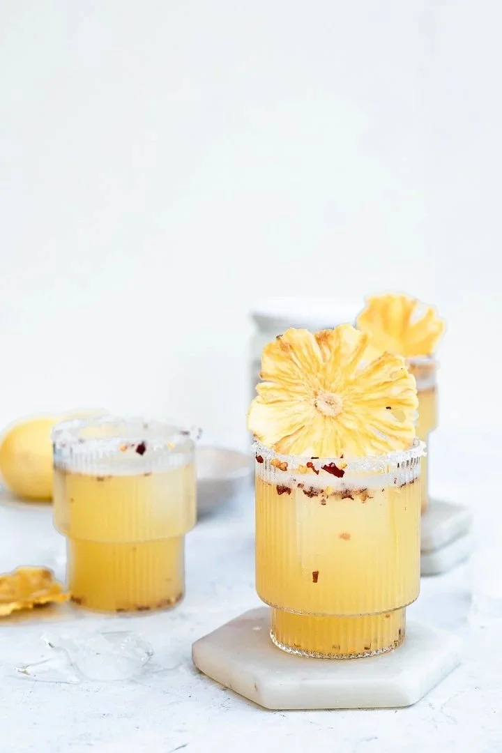 Three glasses of yellow beverage garnished with dried pineapple slices, on a white surface with ice and lime in the background.