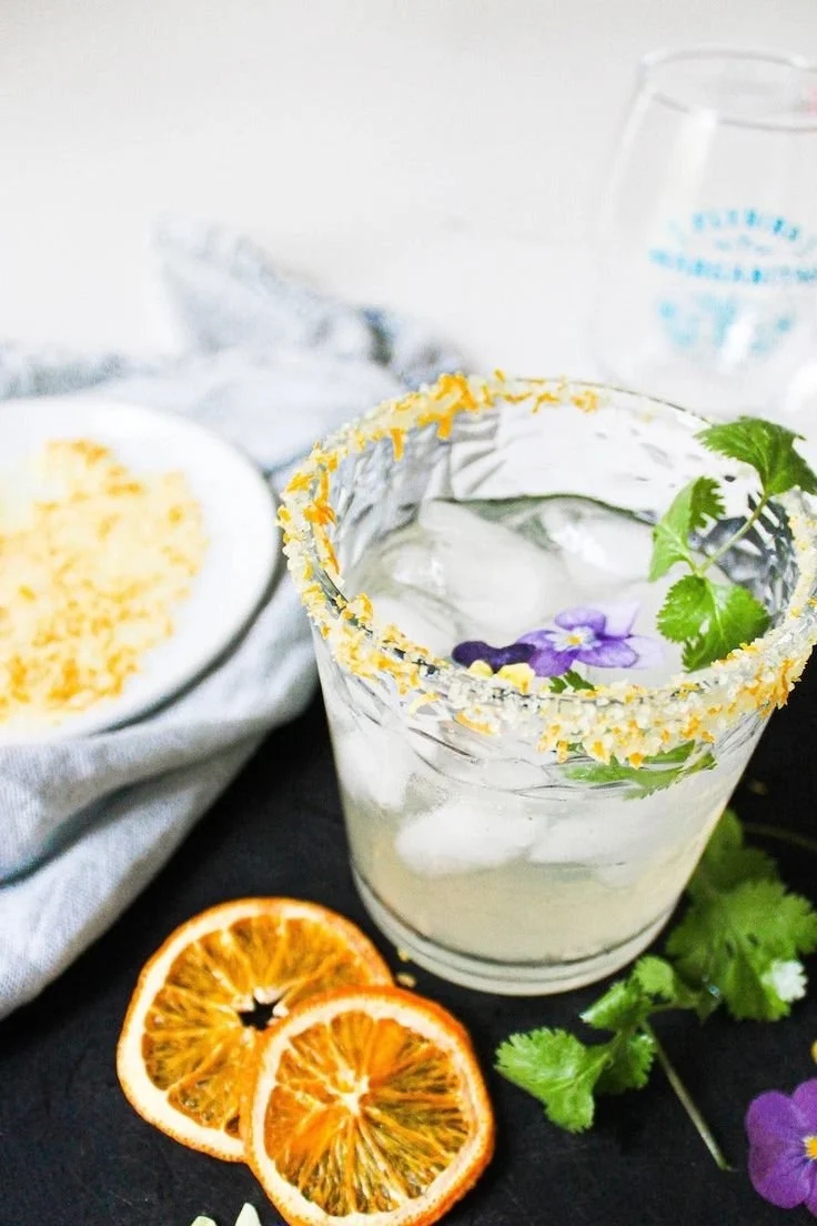A glass of ice water garnished with a slice of lime, a sprig of cilantro, purple flowers, and a salted rim, placed on a dark surface with dried citrus slices nearby.