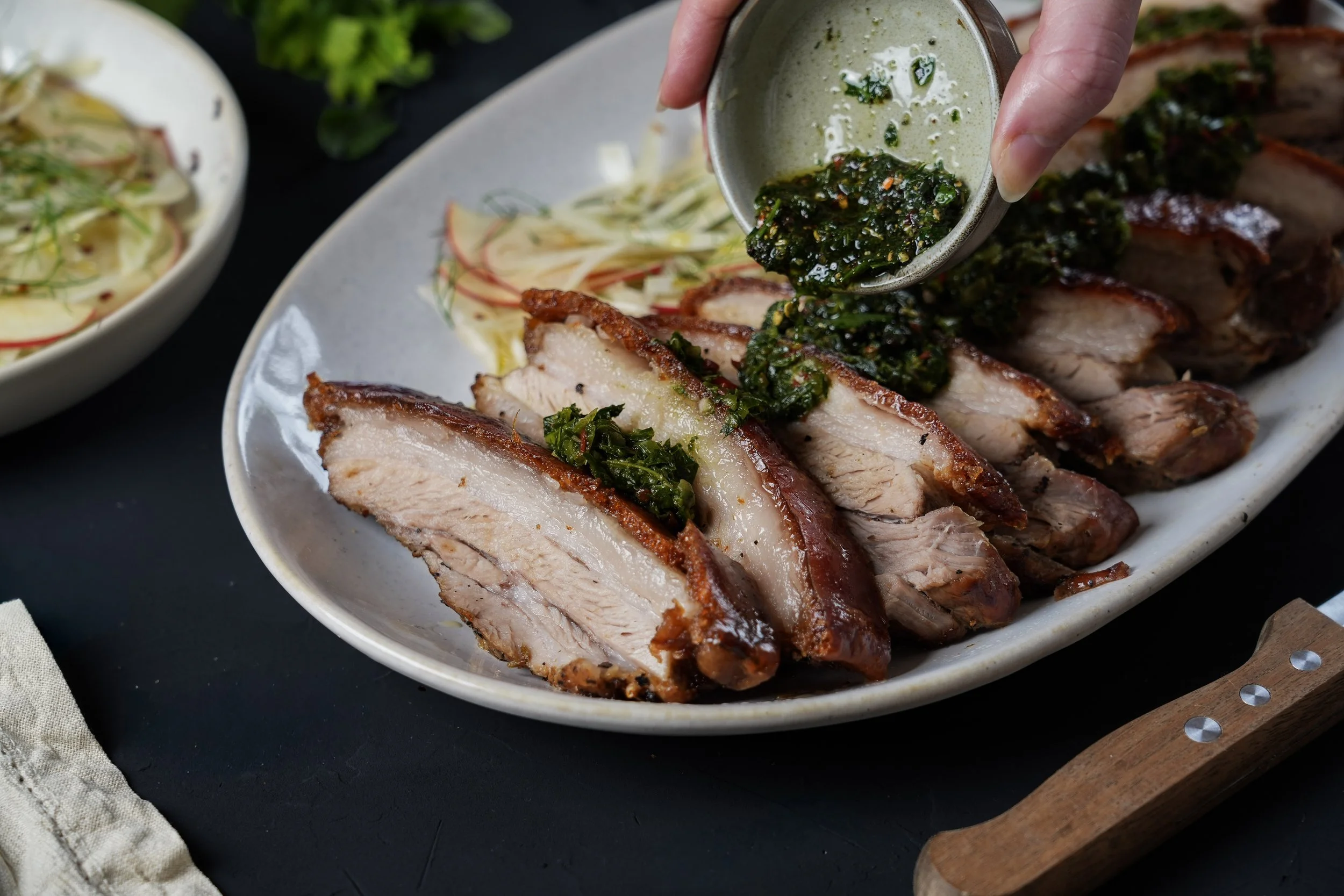 Sliced roasted pork topped with green herb sauce on a white platter, with a bowl of salad and a wooden serving board nearby.