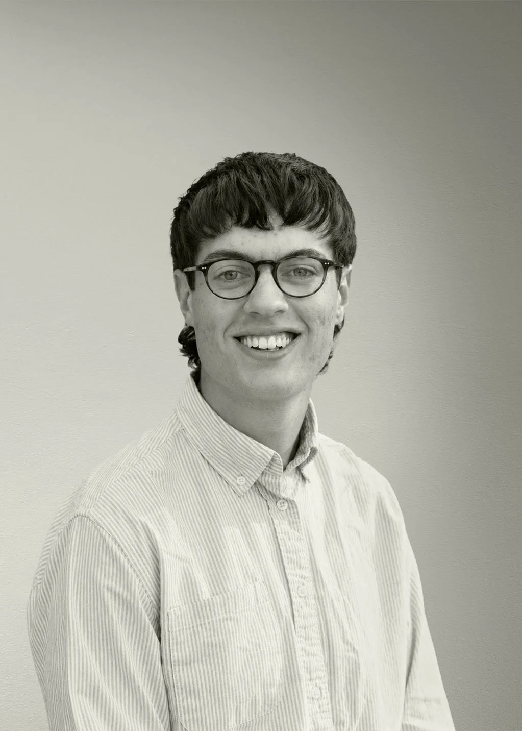 Black and white portrait of a young man with dark hair, glasses, and a striped button-up shirt, smiling and standing against a plain background.