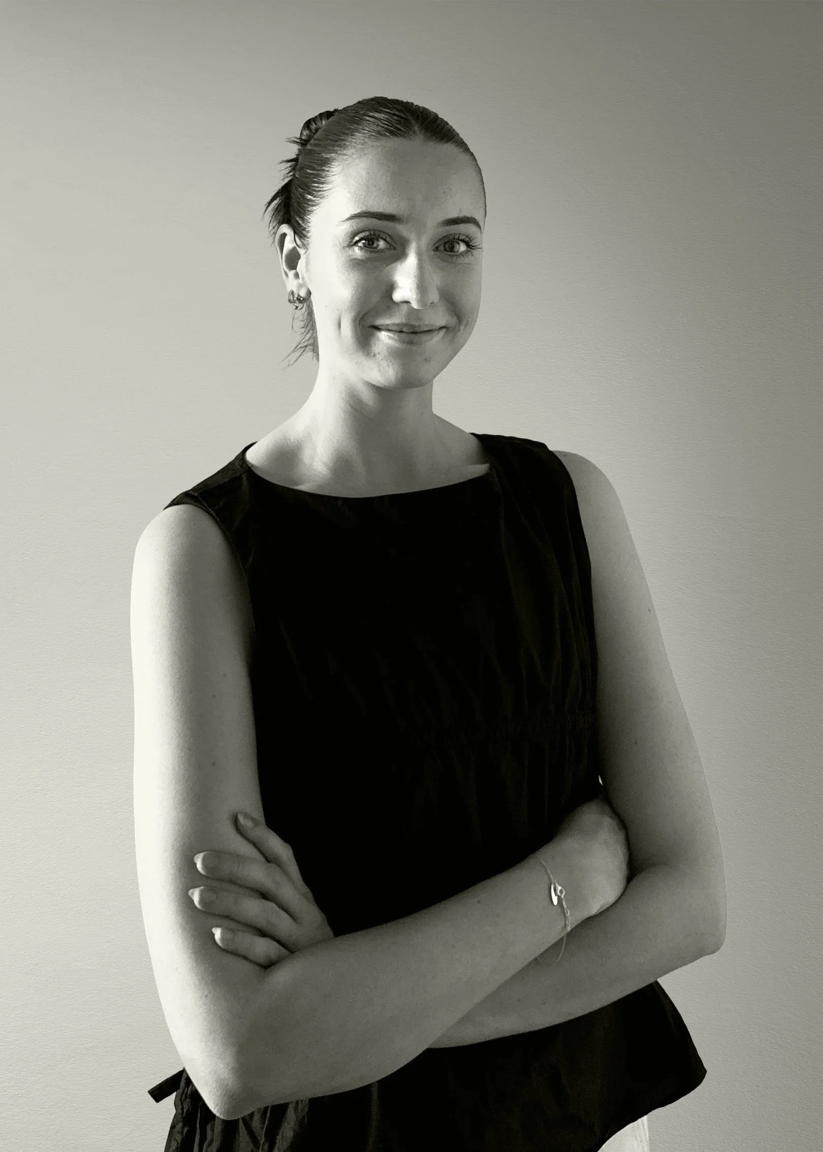 Black and white photo of a woman with her arms crossed, smiling, wearing a sleeveless top and earrings, standing against a plain light-colored wall.