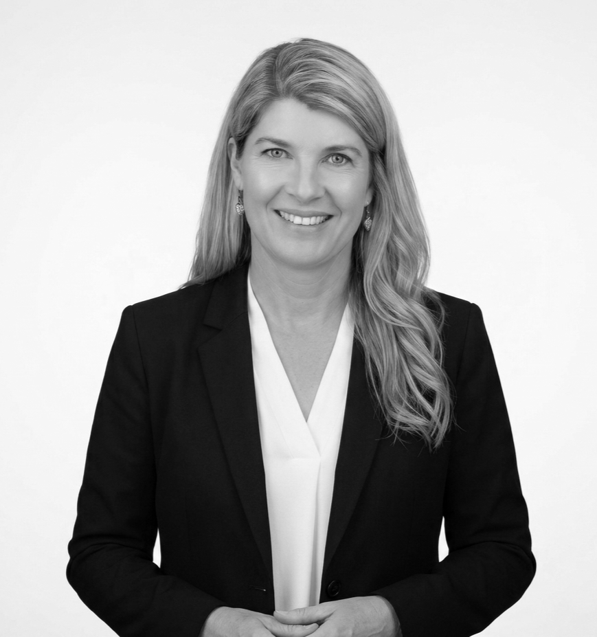 Black-and-white portrait of a smiling woman with long wavy blonde hair, wearing a black blazer over a white blouse, against a plain light background.