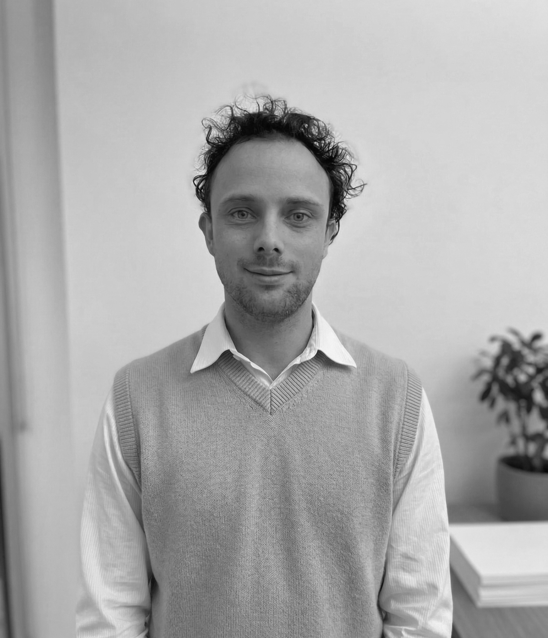 A man with curly hair and light eyes standing indoors, wearing a white collared shirt and a light gray sweater vest, in front of a plain wall with a plant and a table in the background.