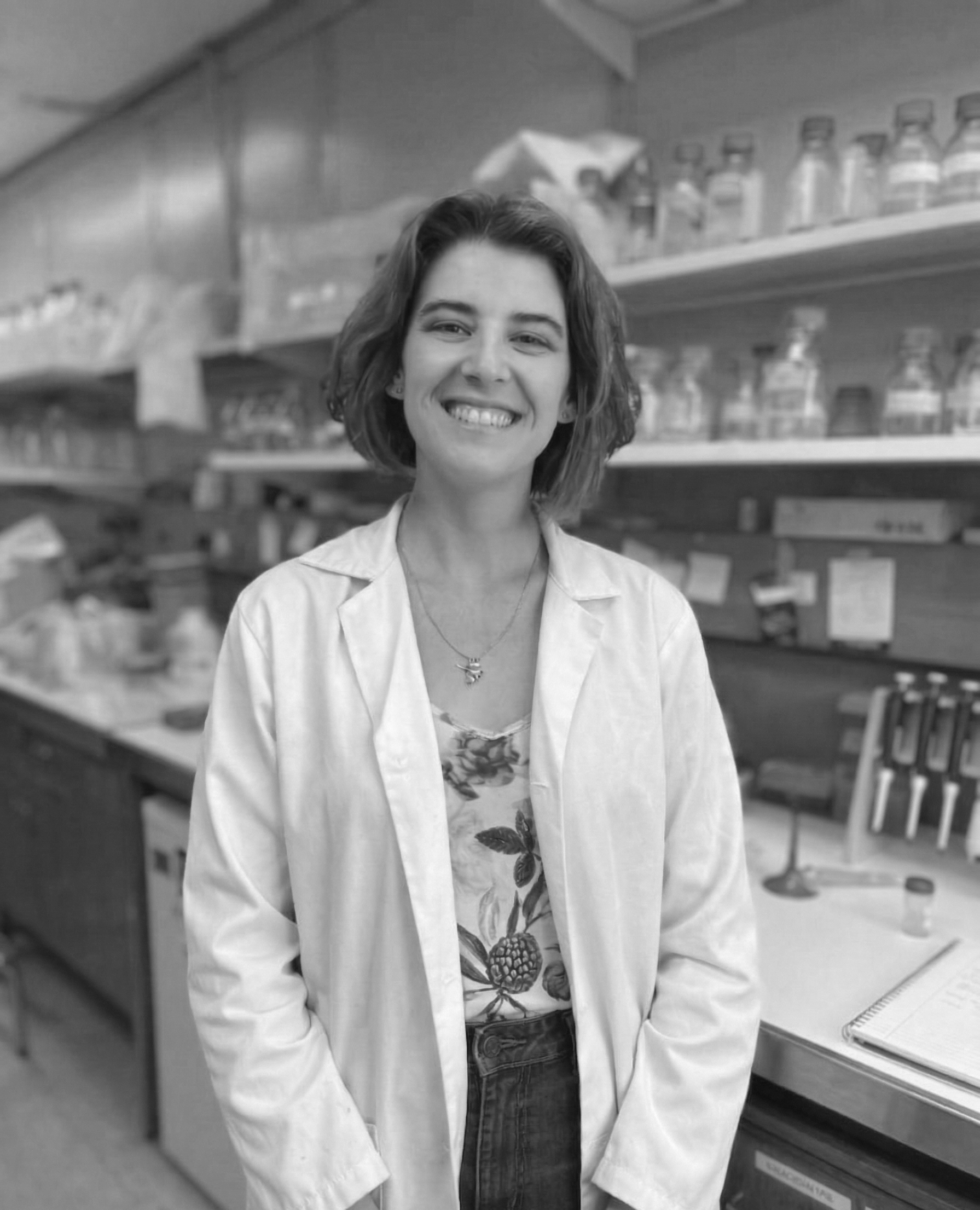 A woman with short, wavy hair smiling in a laboratory or pharmacy setting, wearing a white lab coat over a floral blouse with shelves of jars and laboratory equipment in the background.