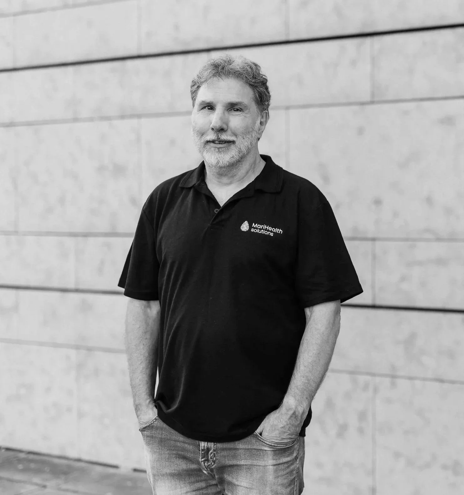 A middle-aged man with curly hair and a beard standing outdoors against a stone wall. He is wearing a black polo shirt with a logo and text that reads 'ManiHealth solutions,' and has his hands in his pockets.