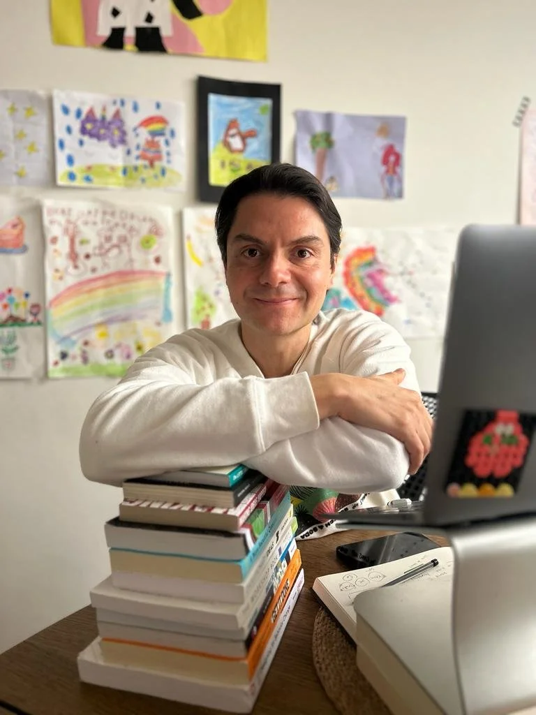 A man sits at a desk with a stack of books, smiling at the camera. Behind him on the wall are colorful drawings and paintings, some featuring rainbows, animals, and figures.