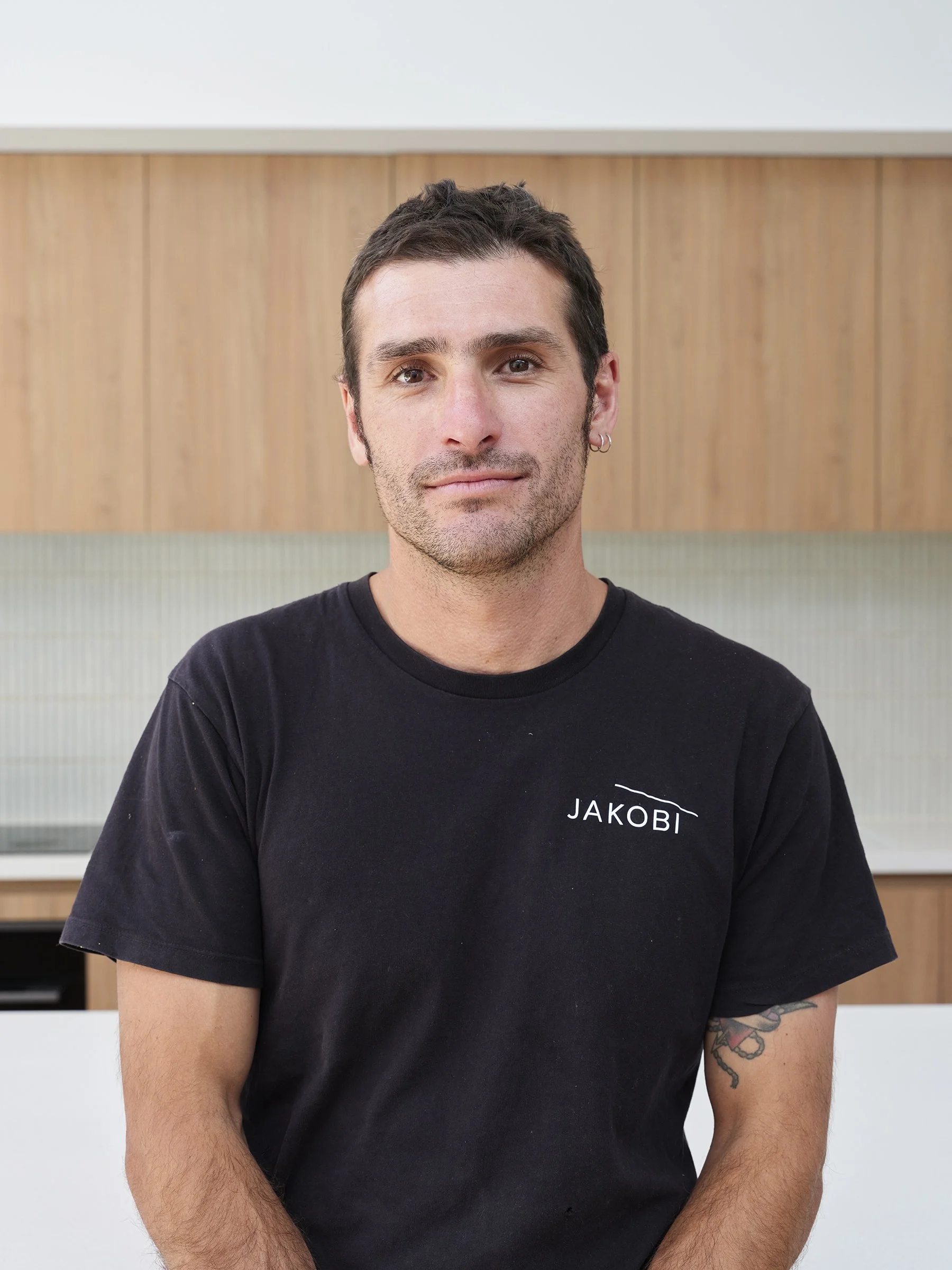 A young man with dark hair, light skin, and earrings, wearing a black t-shirt with "JAKOBI" written on it, standing in front of a background with wood paneling and light-colored walls.
