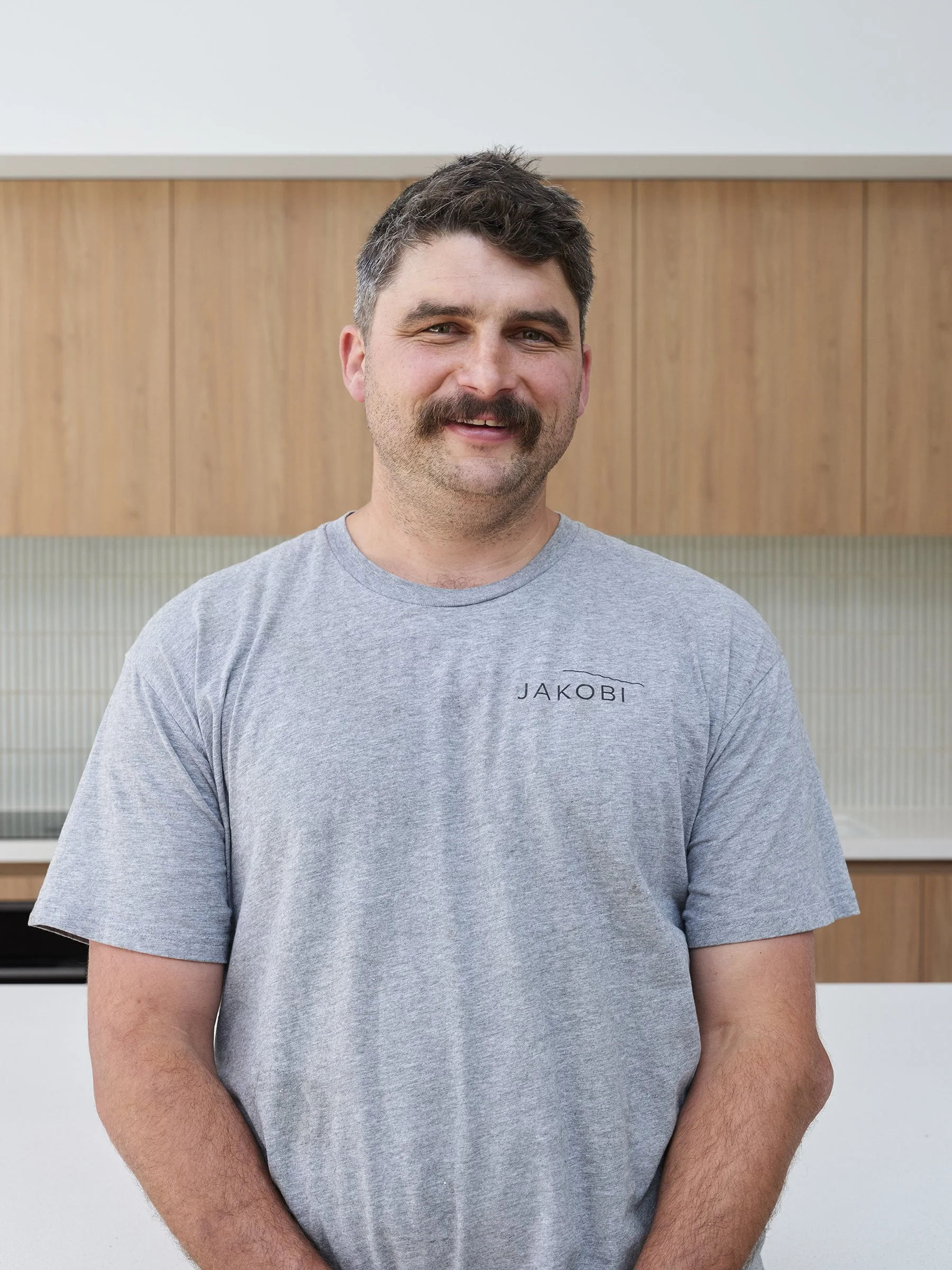 A man with dark hair, a full mustache, and a beard, standing indoors in front of a wooden wall with a glass panel. He is wearing a gray t-shirt with the word 'JAKOBI' printed on it.