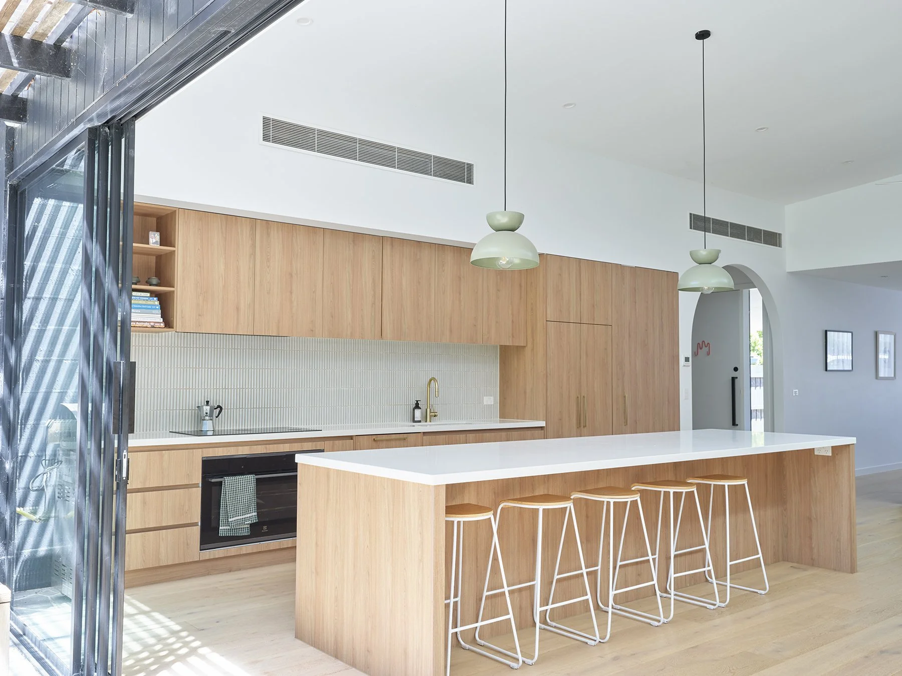 Modern kitchen with wooden cabinetry, white island counter with four white bar stools, pendant lights, built-in oven, and minimalist decor.