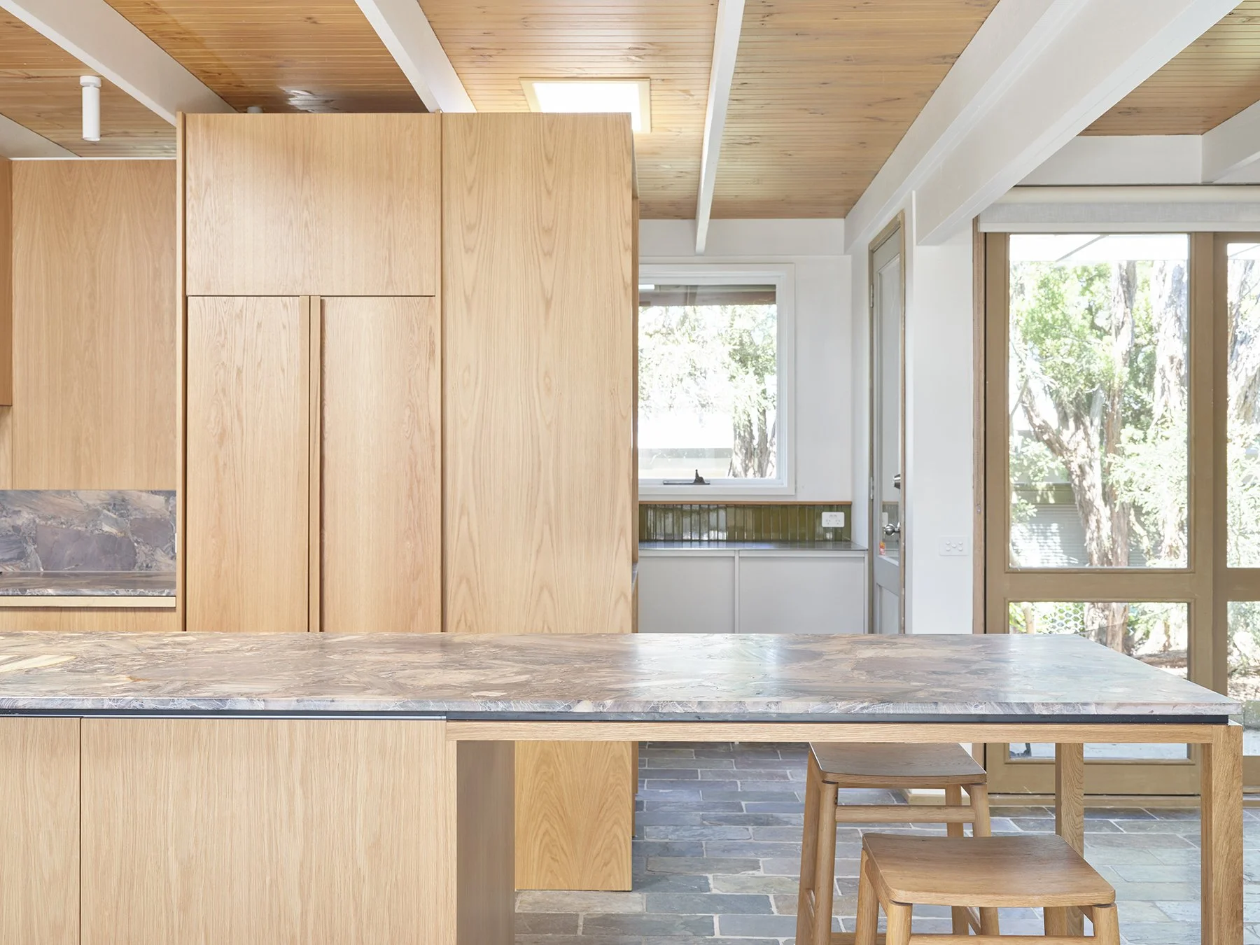 Modern kitchen with wooden cabinets, marble countertop, large windows showing trees outside, and a breakfast bar with wooden stools.
