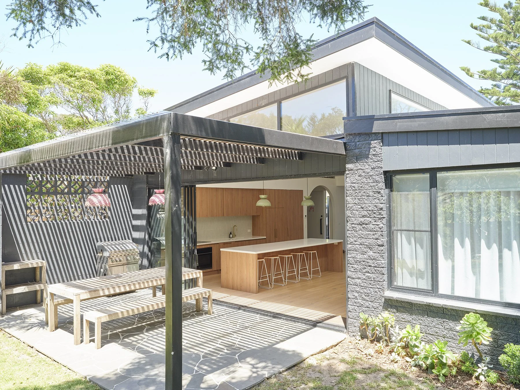 Modern house with outdoor patio area shaded by a pergola, featuring a wooden bench, a barbecue grill, and a partially visible kitchen with white counter and wooden cabinets, surrounded by greenery.
