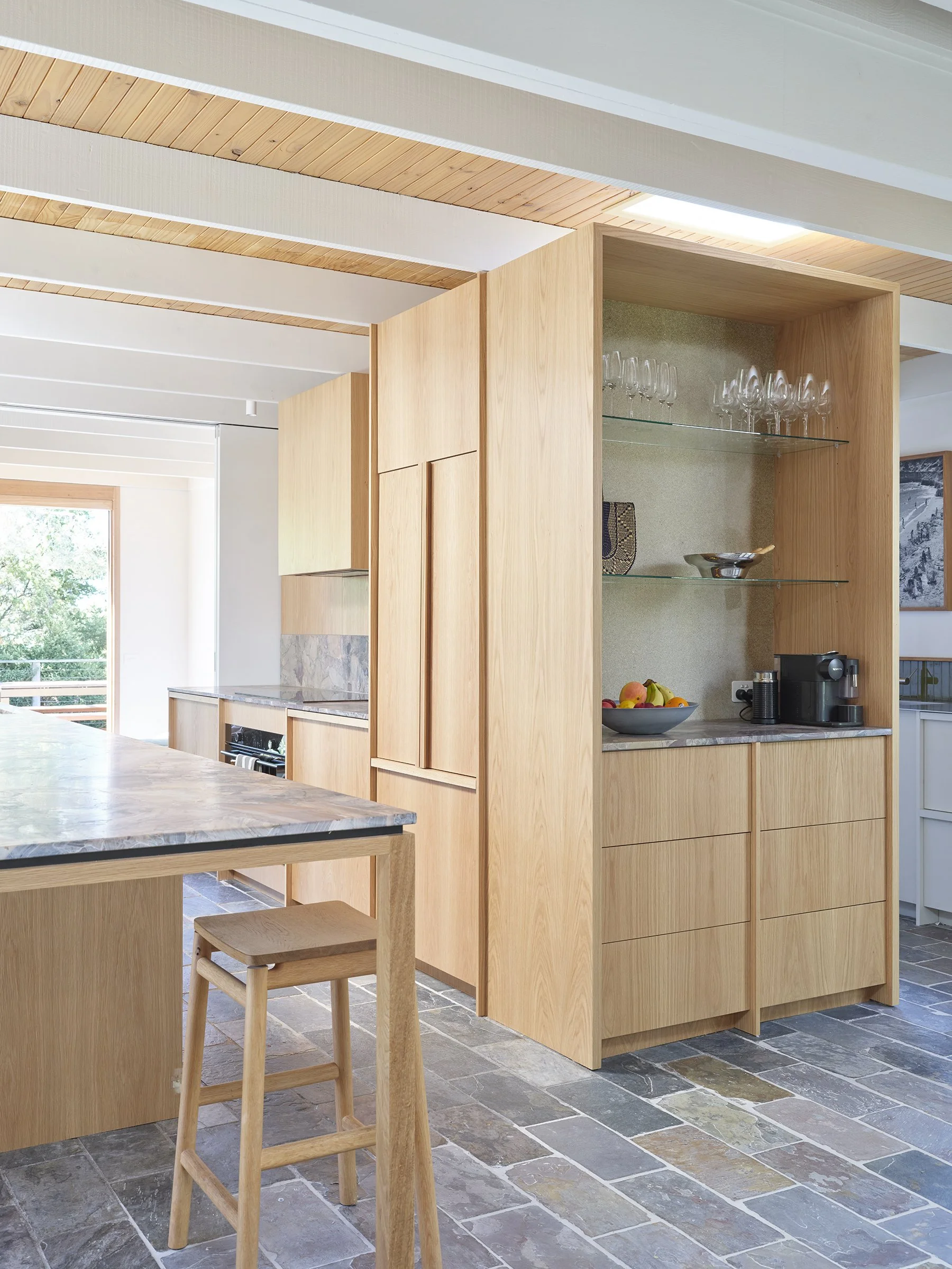 A modern kitchen with light wood cabinets, a marble countertop island, a wooden barstool, a glass cabinet displaying wine glasses, and a window with a view of greenery outside.
