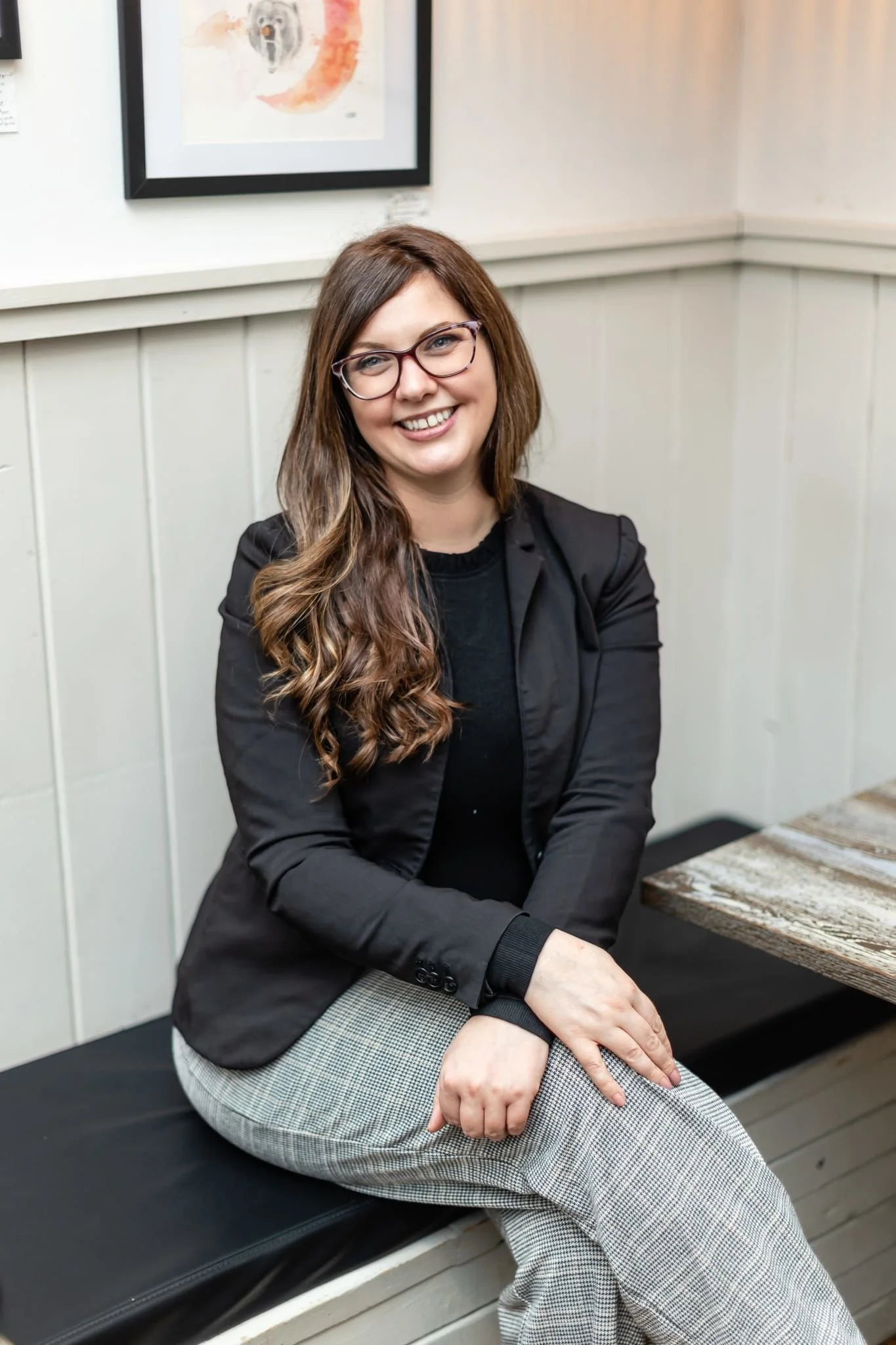 A woman with long brown hair, glasses, and a black jacket, smiling and sitting on a bench indoors. She appears to be at a casual gathering or waiting area, with artwork on the wall behind her and a wooden table nearby.