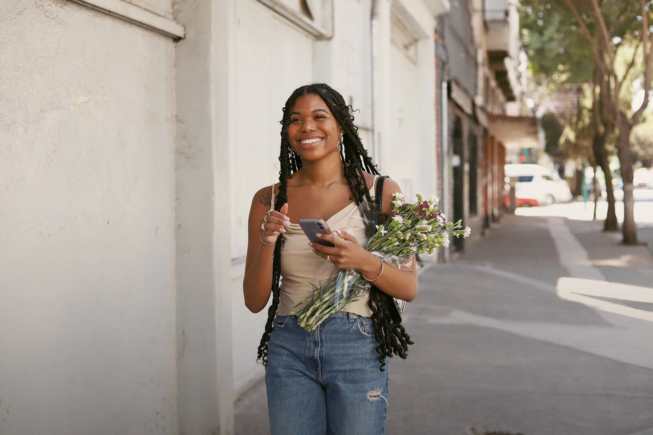 A young woman with long braids smiling while walking on a sidewalk holding a bouquet of flowers and looking at her phone. Created for Canopy, influencer marketing, designed by Asa Rodger at Page®.