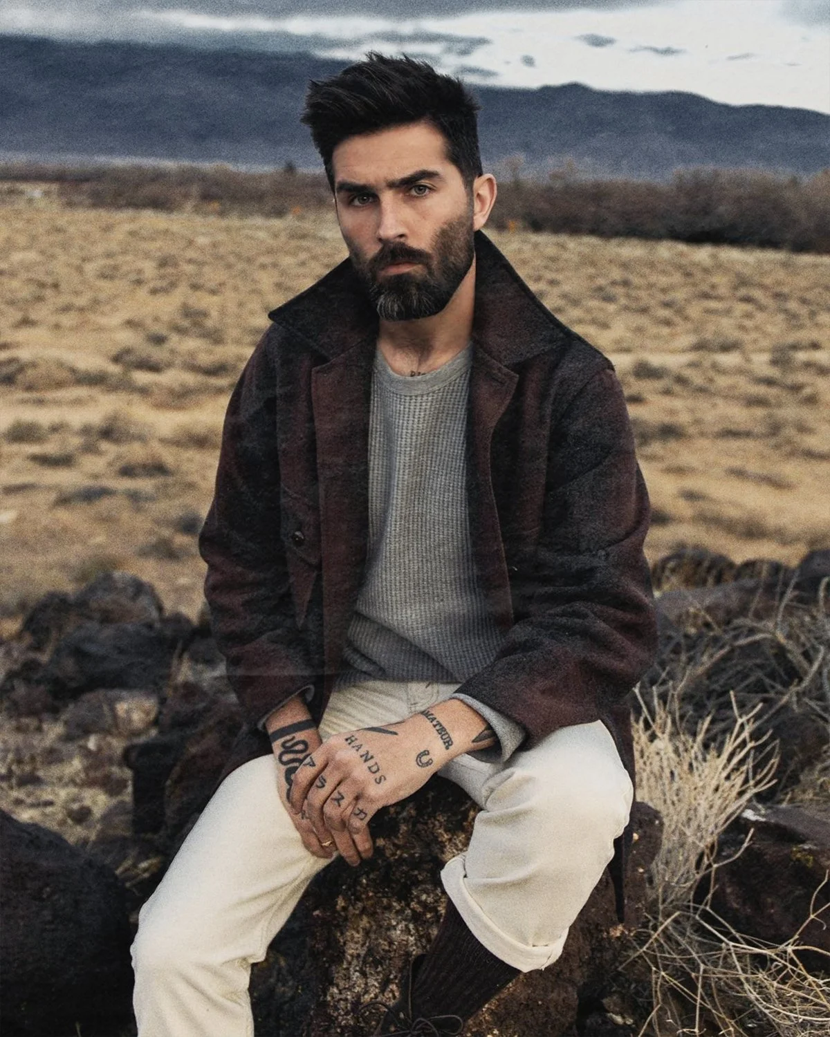 Chris Millington modelling, sitting outdoors in a rocky, desert landscape during cloudy weather, wearing a dark jacket, gray sweater, and light-colored pants.