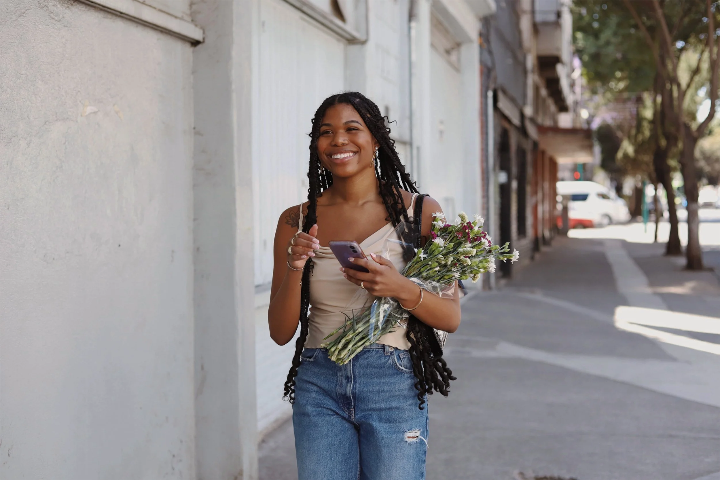 A young woman with long braids smiling while walking on a city sidewalk, holding a bouquet of flowers and a smartphone. Created for Canopy, influencer marketing, designed by Asa Rodger at Page®.