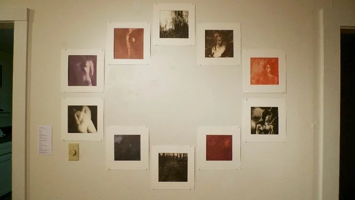 A wall with twelve framed black and white or sepia-toned photographs arranged in a circle, some images depicting nude or semi-nude figures. There is a small paper note on the left side of the wall and a light switch below it. 