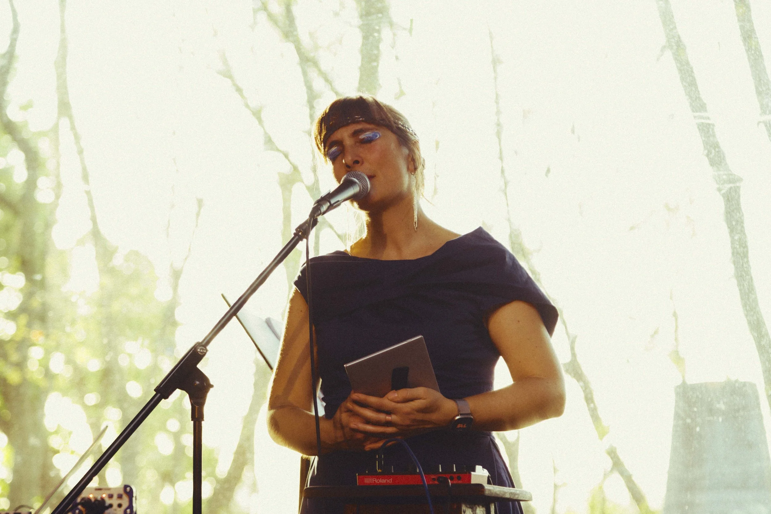 A woman singing into a microphone during a live performance in front of a window with trees outside and dappled sunlight.