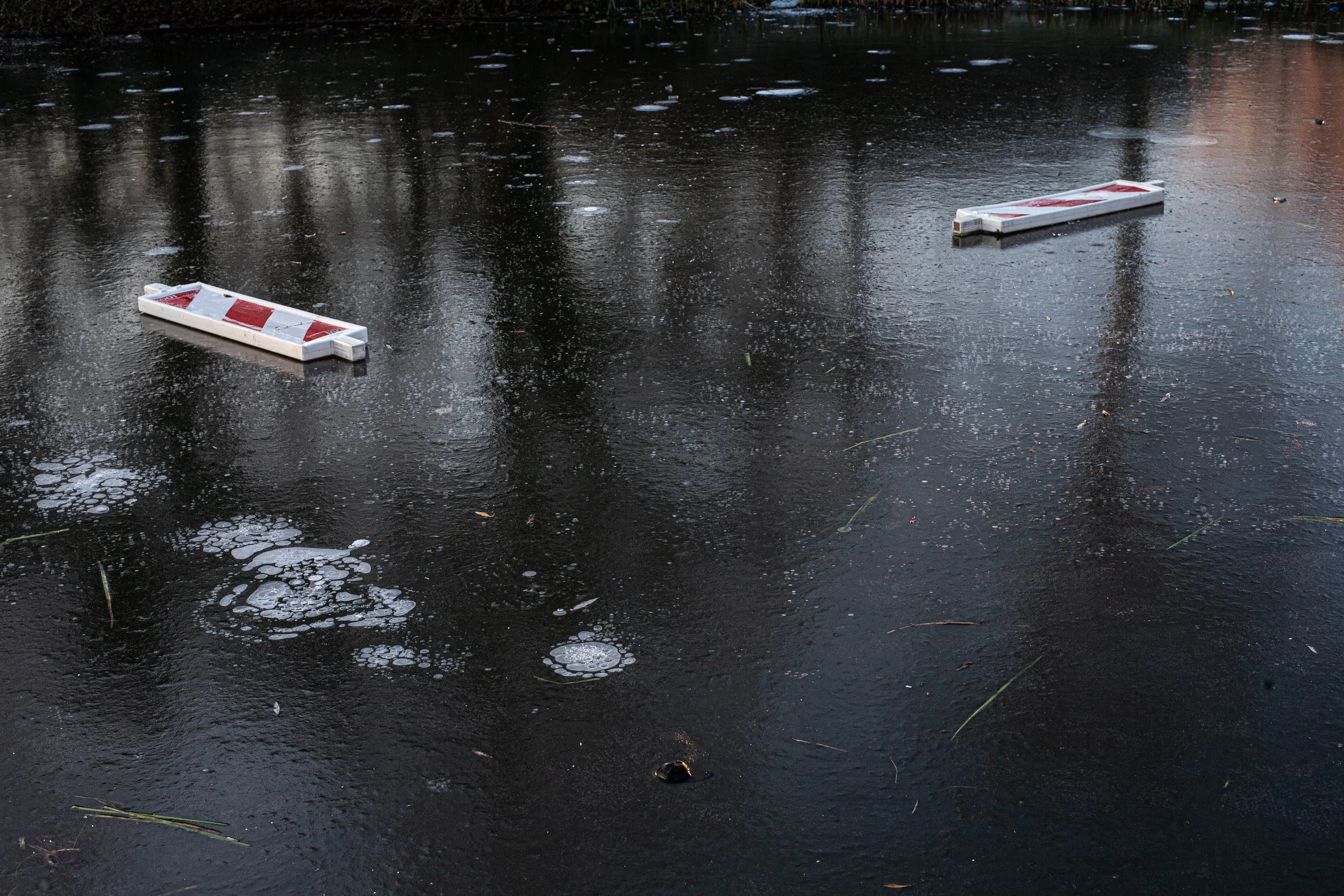 A frozen canal with two fallen traffic barriers, puddles, and ice patches.