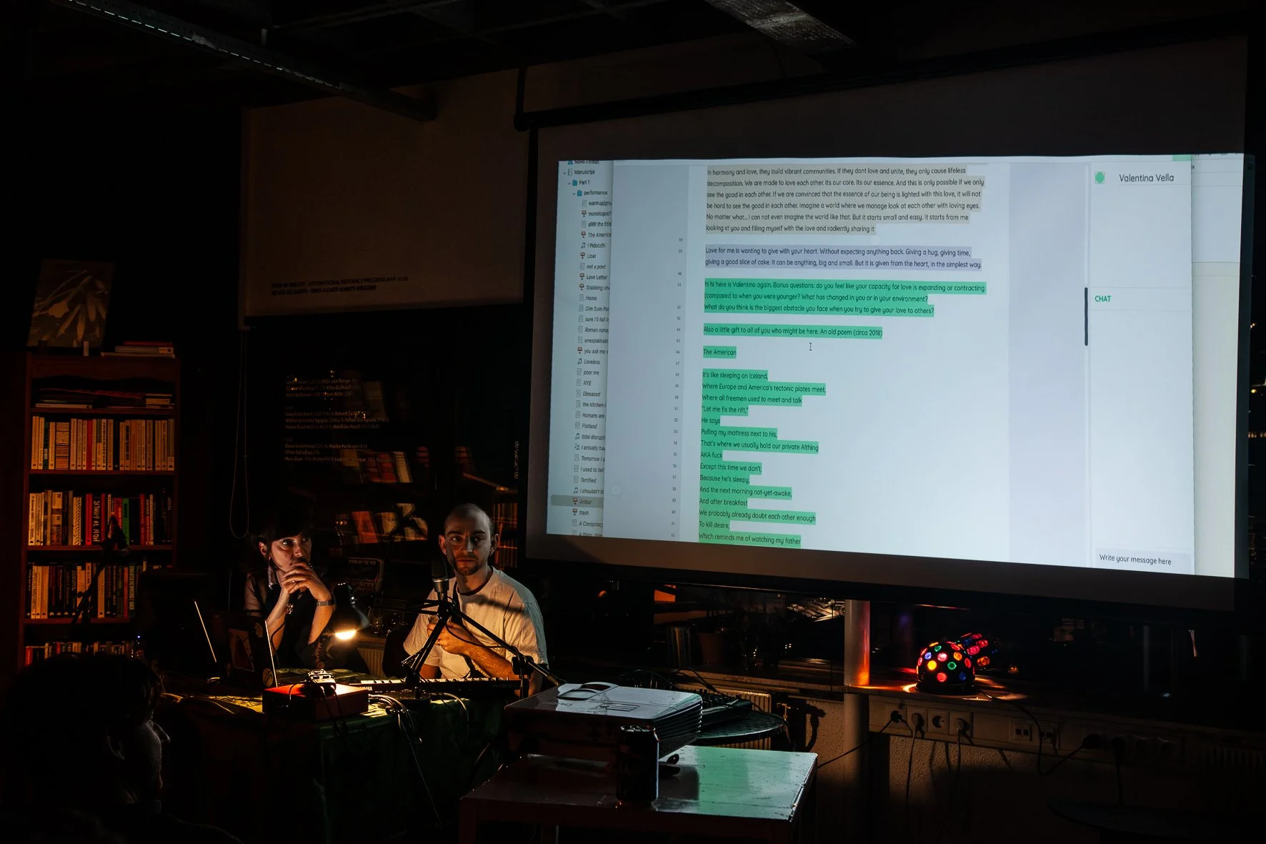A dark room with a large projector screen displaying a chat conversation or script. Two people are sitting at a table in front of microphones and laptop, with bookshelves on the left and a colorful light ball on the right.
