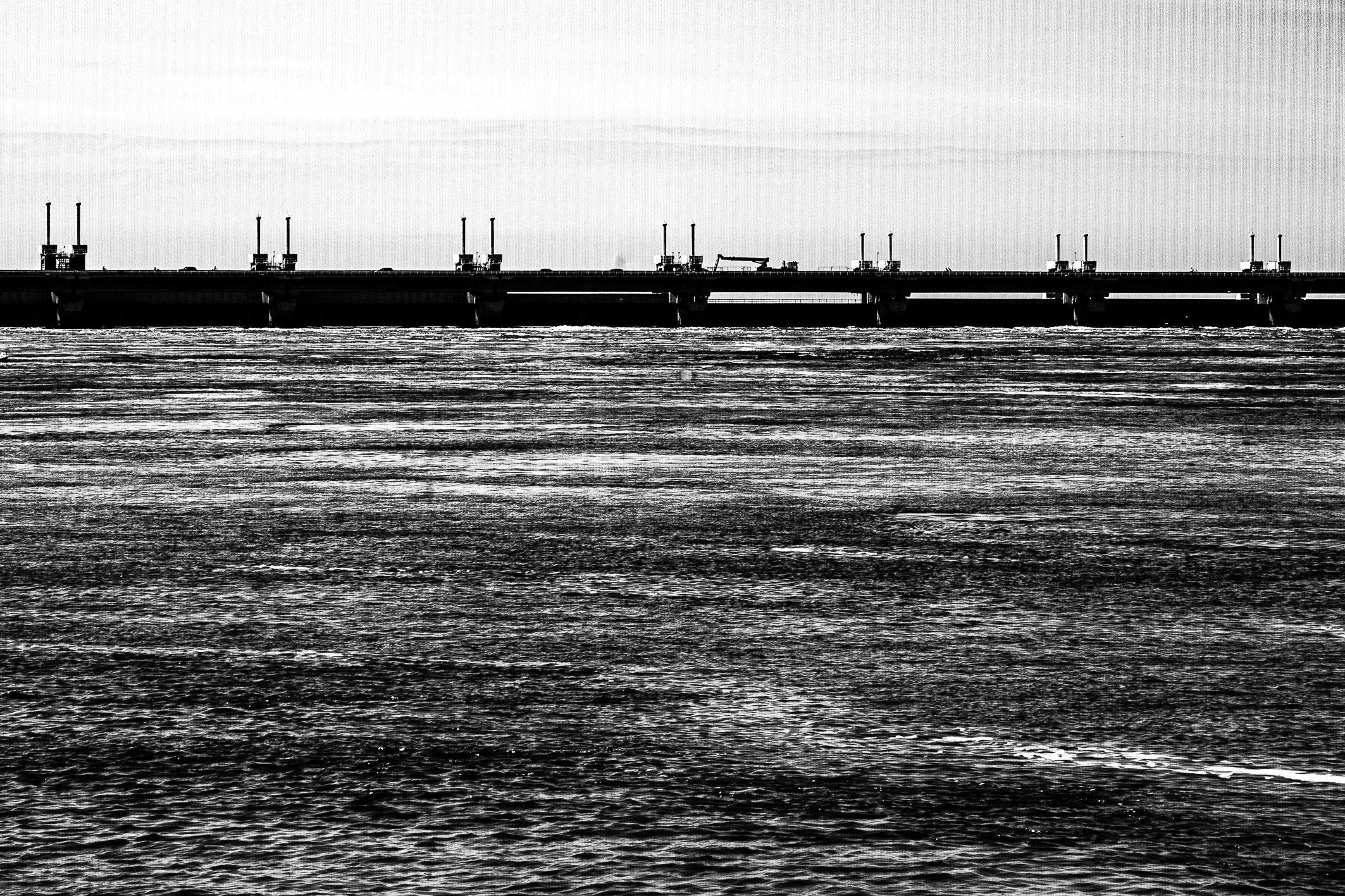 Black and white photo of a long pier extending into the water with multiple cranes or hoists along it, against a cloudy sky.
