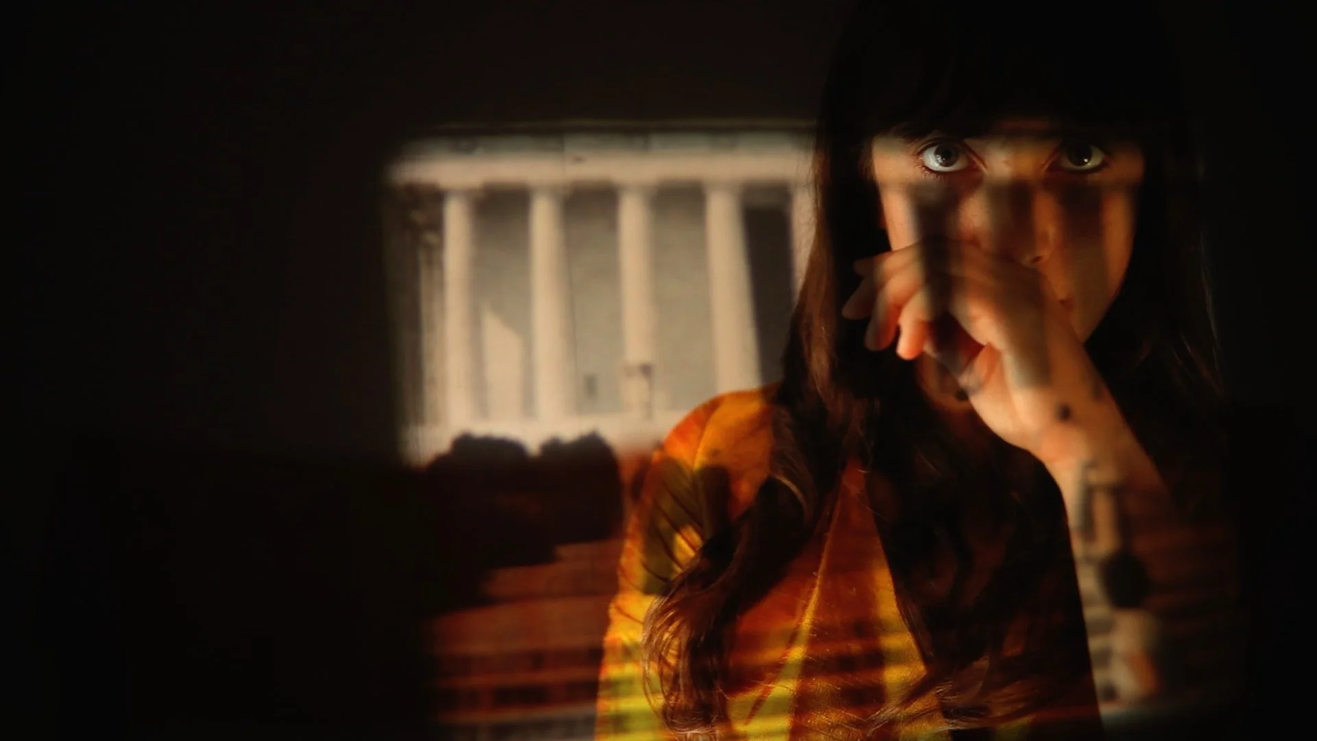 A woman with brown hair and light skin with her hand covering her mouth, in a dark setting. The background shows blurry architecture from Washington DC.