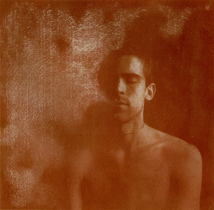 A shirtless young man with curly hair, standing next to a cloud of smoke with a contemplative expression.