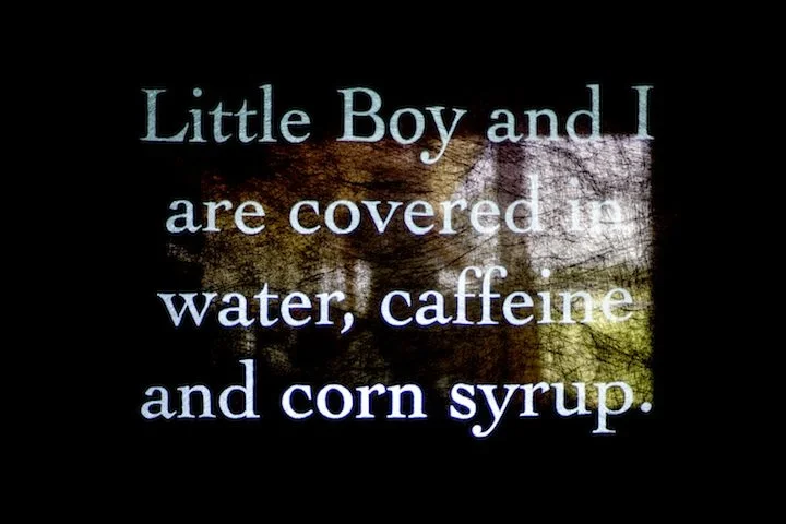projected text on a scrim reads: Little Boy and I are covered in water, caffeine and corn syrup