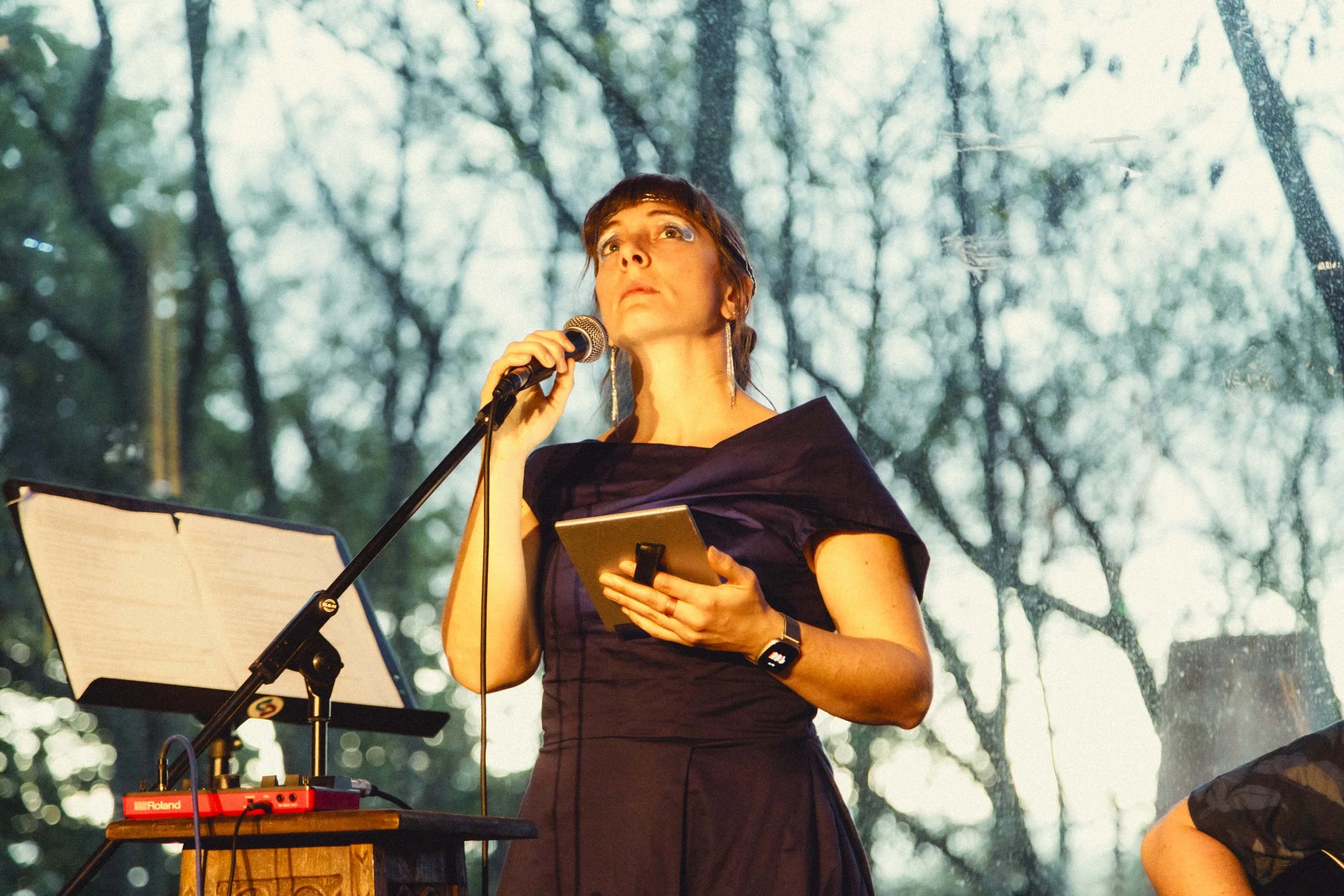 A woman with short dark hair and earrings, dressed in a black outfit, holding a microphone and a tablet, standing on an outdoor stage with a music stand and equipment, with trees and a cloudy sky in the background.